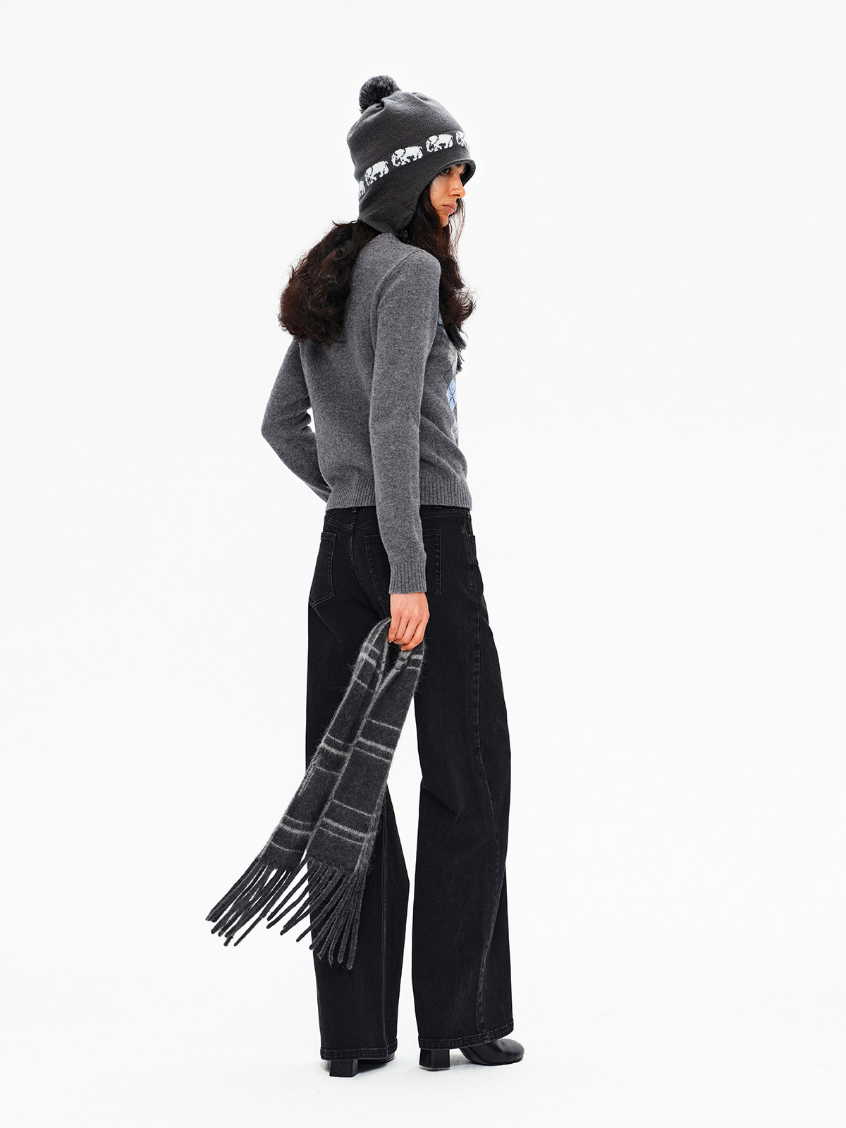 A woman in a gray sweater, black wide-leg pants, and a beanie stands sideways holding the EastLyst Karl alpaca wool classic scarf. The minimalist design is highlighted by a plain white background.