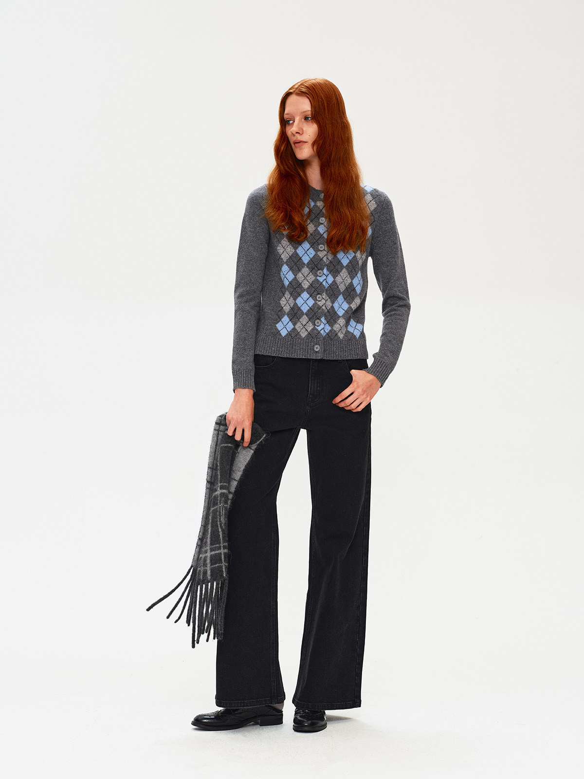 A woman with long red hair wears a gray argyle sweater, black wide-leg pants, and black shoes. She holds the EastLyst Karl alpaca wool classic scarf and stands against a plain white background, expressing minimalist style.