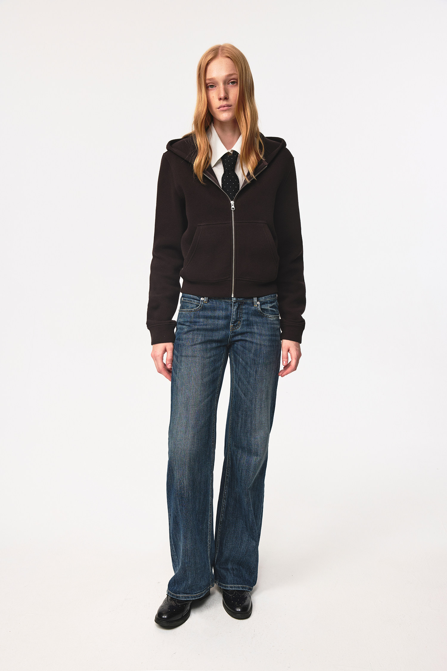 A person with long blonde hair wears a black zip-up hoodie, white shirt, black tie, EastLyst Slub Textured Casual Pants in indigo wash, and black shoes, standing against a plain white background.