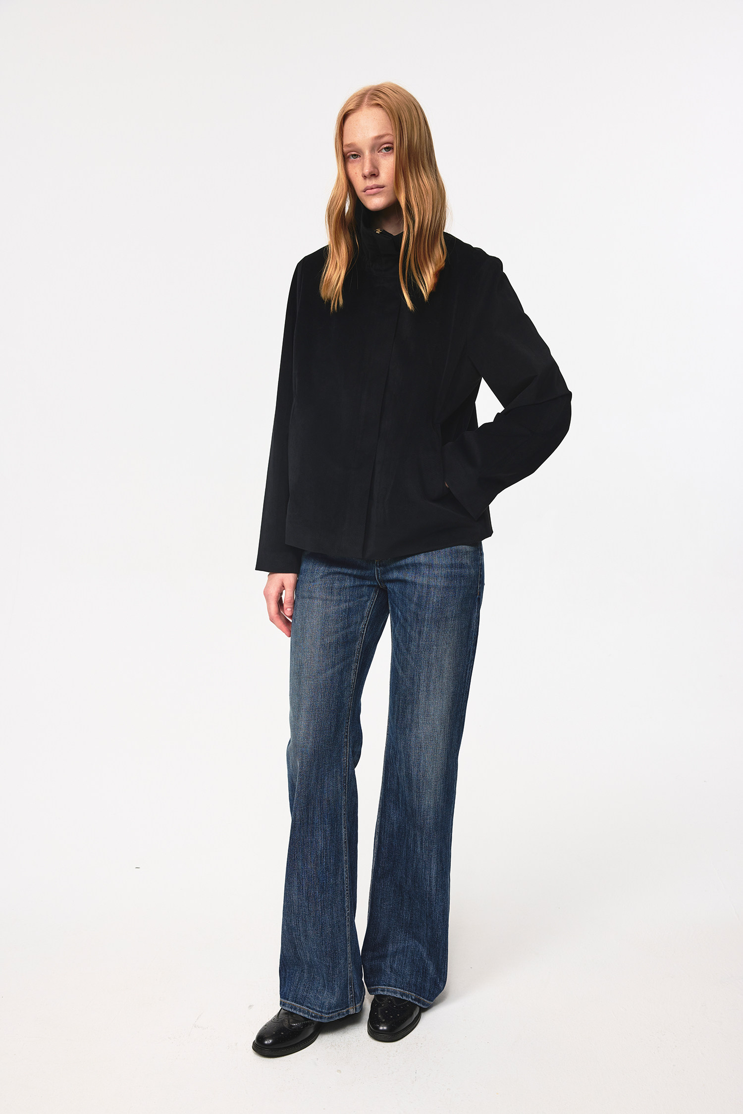 A person with long blonde hair stands against a white background, wearing a black jacket, EastLyst Slub Textured Casual Pants in vintage indigo wash, and black shoes, with a neutral expression and one hand in their pocket.