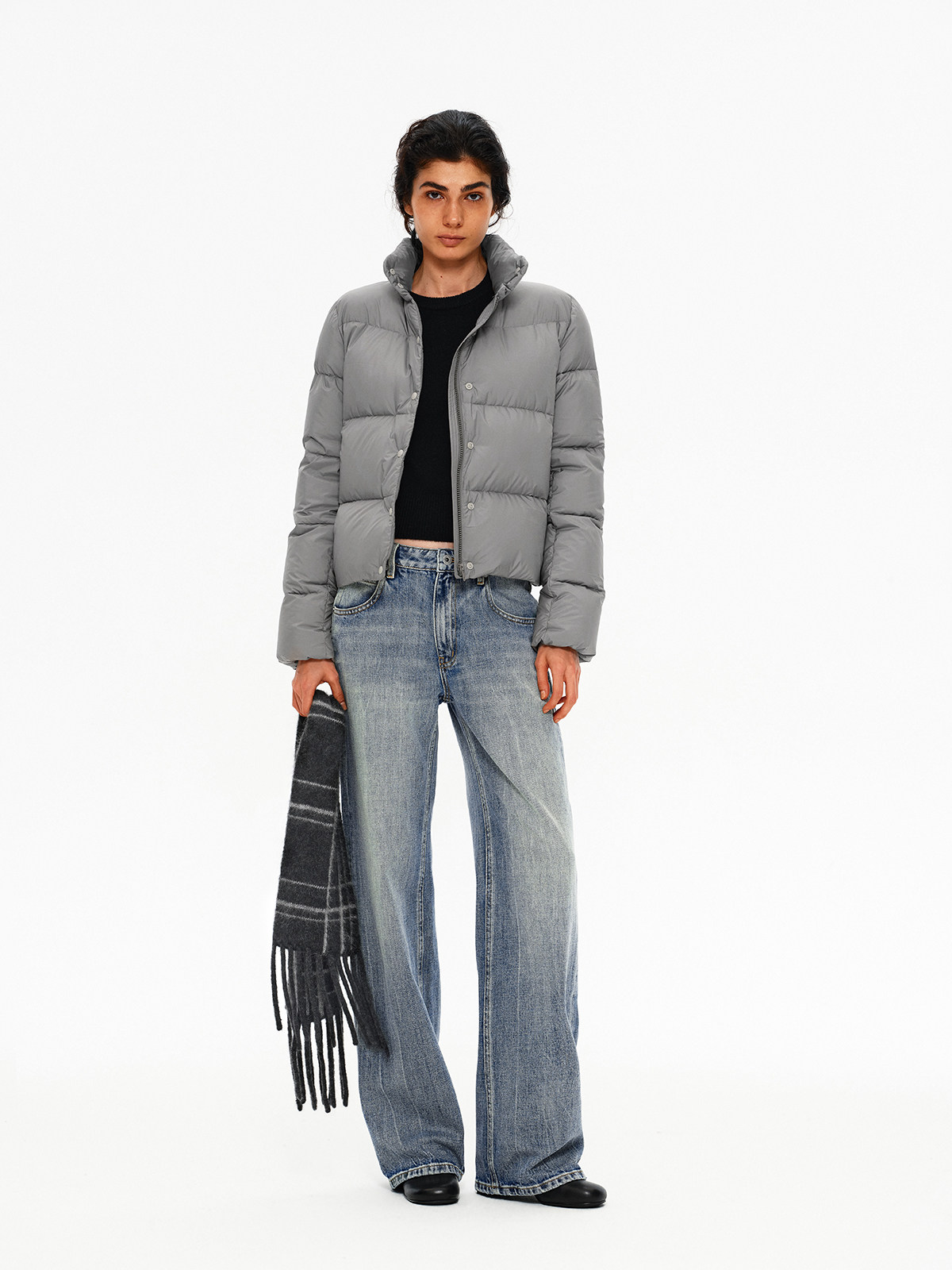A person stands against a white background in a gray puffer jacket, black top, wide-leg blue jeans, and black shoes, holding the EastLyst Karl alpaca wool classic scarf in their left hand.