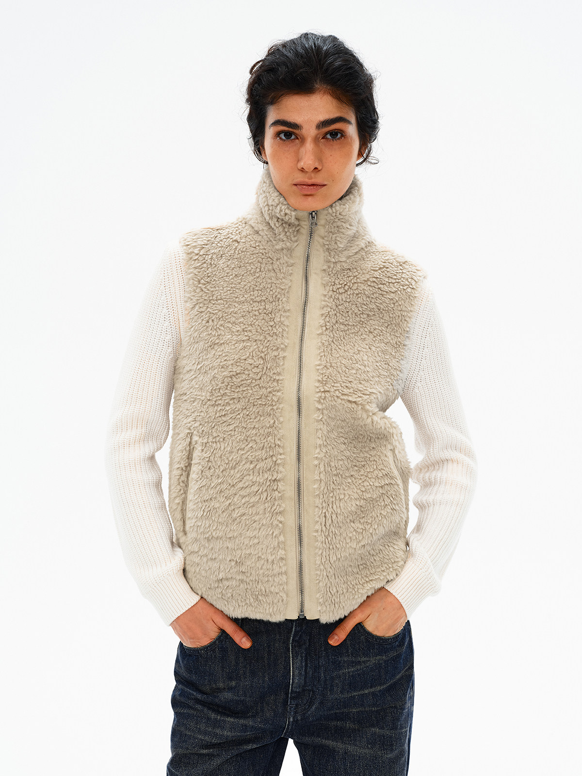 一位黑发模特身穿 EastLyst Curly Sherpa Gilet with High Neck（一款舒适的奶油色冬季背心），内搭白色罗纹毛衣，下身搭配深色牛仔裤，背景为纯白色。