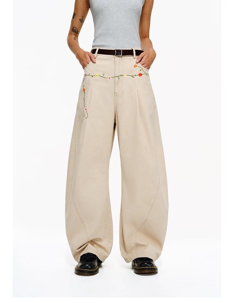 A person stands hands-in-pockets wearing a gray sleeveless top, EastLyst Floral Embroidery Cargo Pants in beige with a string of colorful lights at the waist, black shoes, and a dark belt.