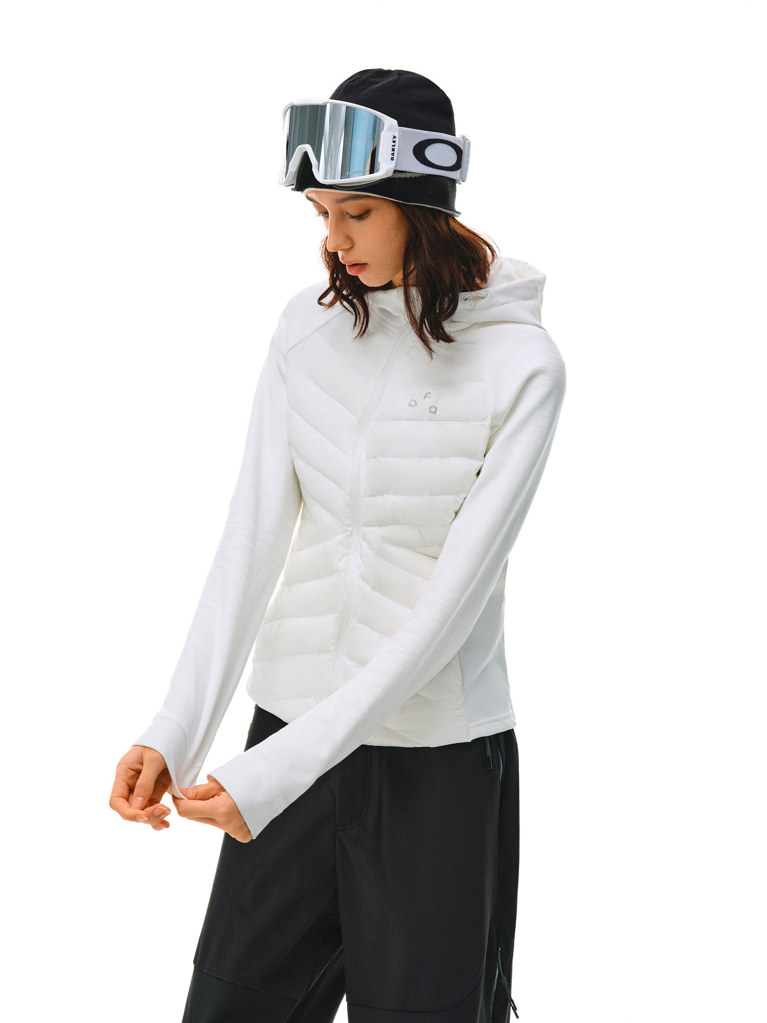 A woman in an EastLyst Quilted Hooded Down Jacket, black pants, and ski goggles stands against a white background, looking down and adjusting her sleeve for lightweight warmth.