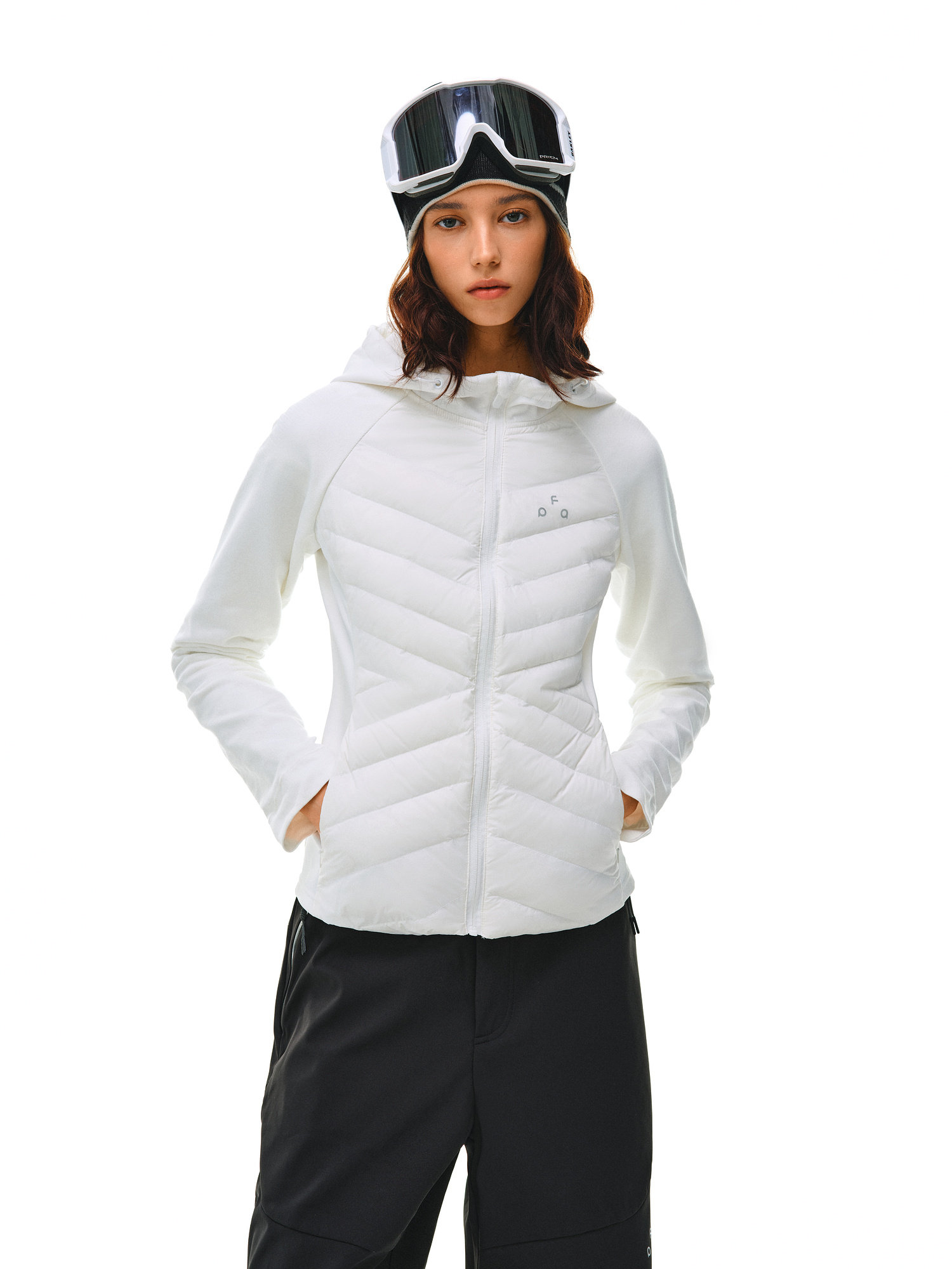 A woman stands facing forward with her hands in her pockets, wearing the EastLyst Quilted Hooded Down Jacket in white, paired with black ski goggles and black pants against a plain white background.