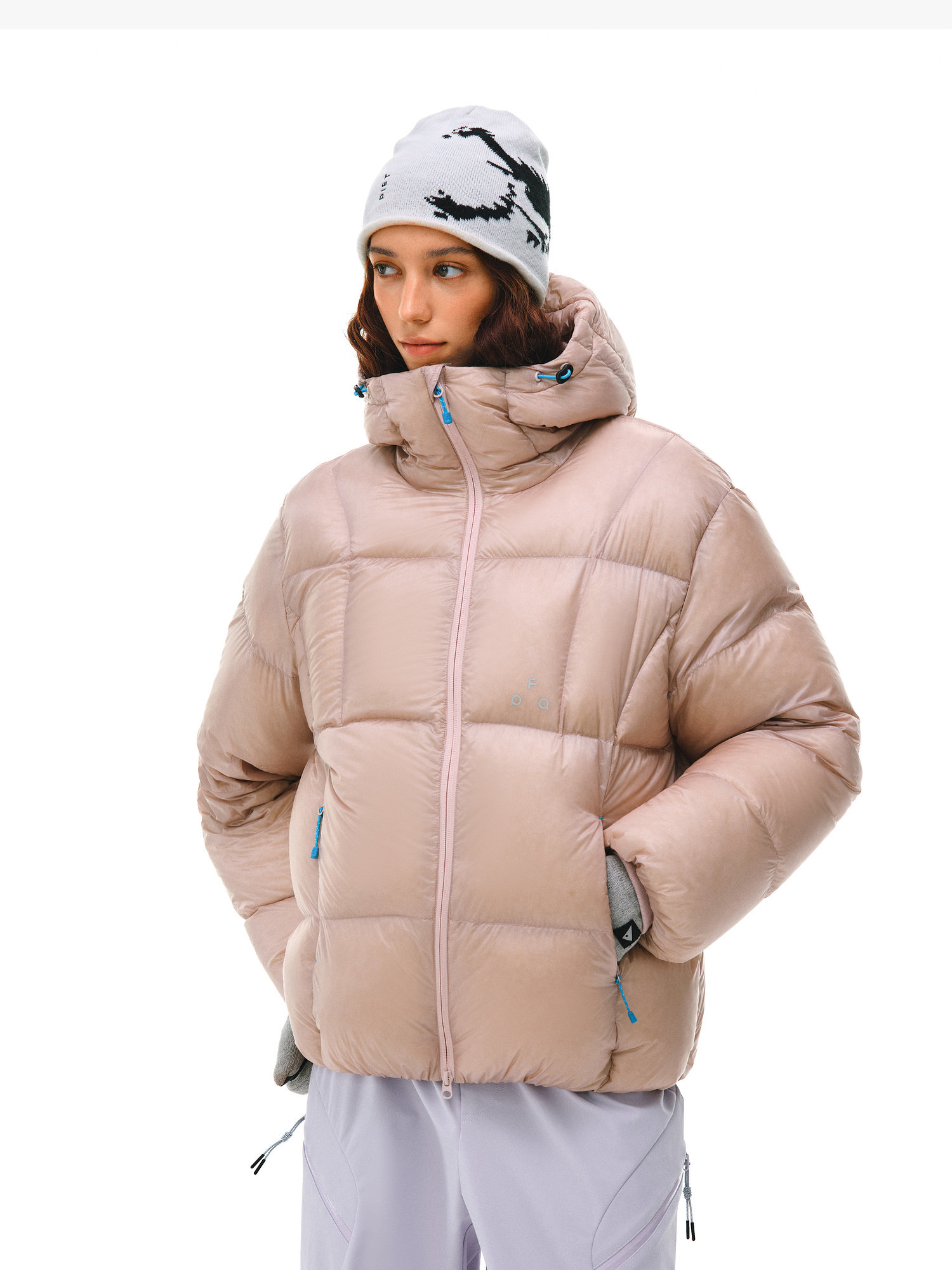 A woman wears the EastLyst Extreme-Cold Down Jacket in light pink, paired with light purple pants and a white beanie with a black graphic, standing hands-in-pockets against a plain white background.