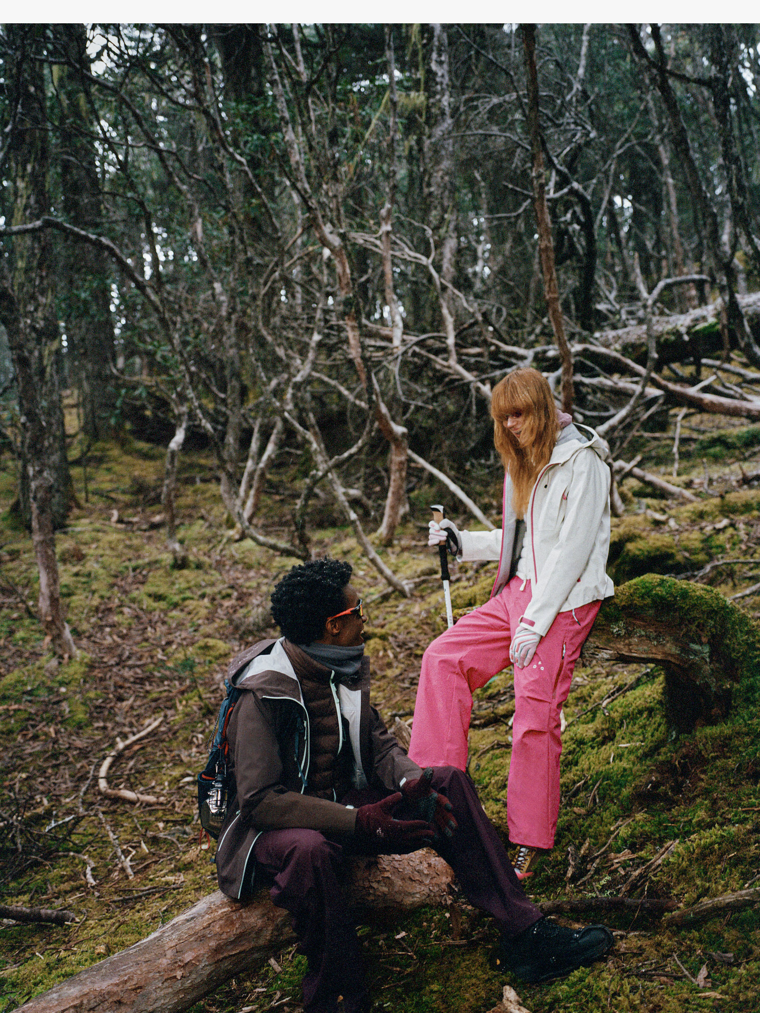 Two people in outdoor gear, including the EastLyst 3-in-1 Color-Block Storm Jacket, explore a mossy forest. One sits on a fallen tree while the other stands with a camera, surrounded by bare branches, highlighting the jacket’s all-weather protection.