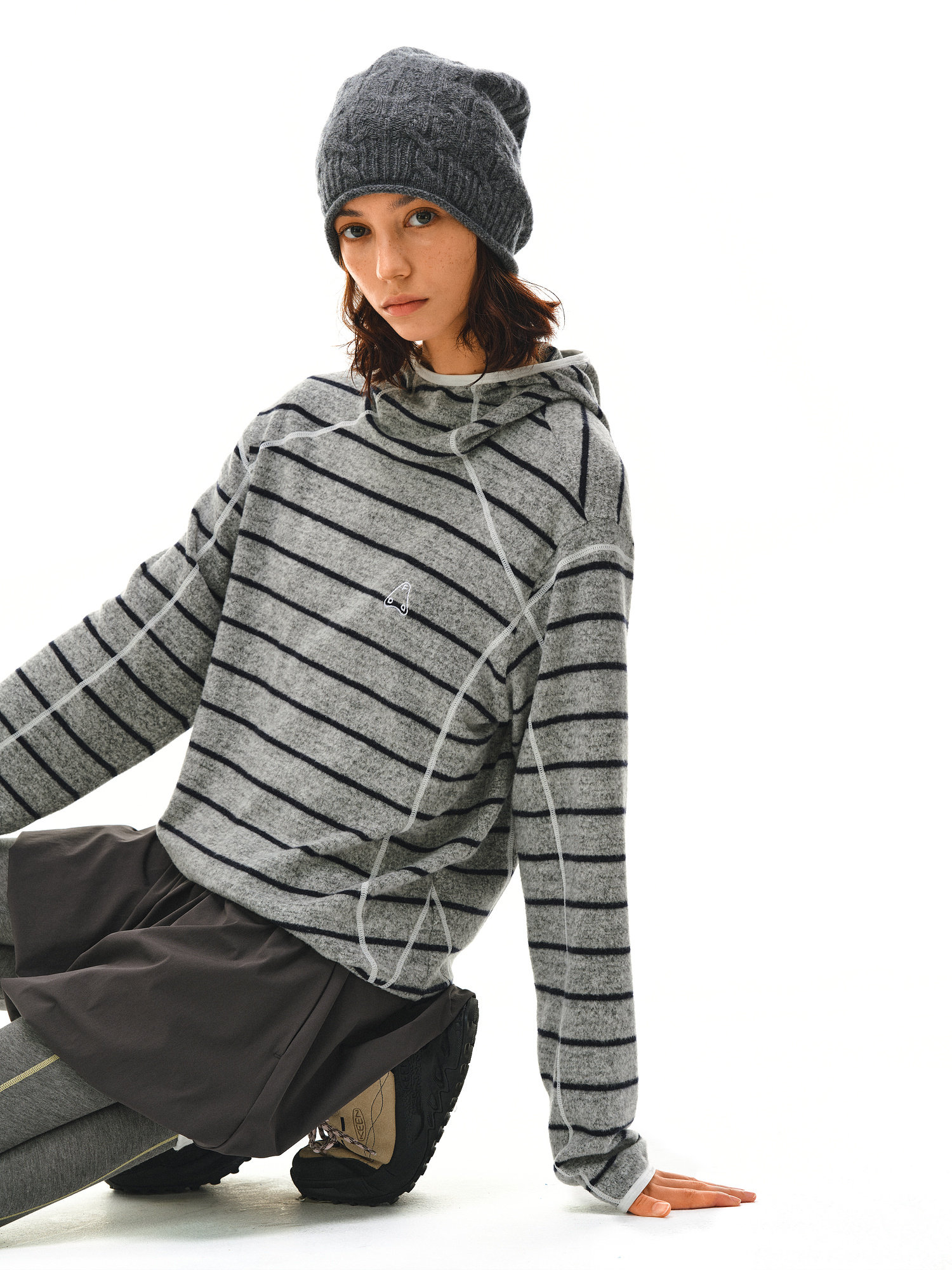 A young person in an EastLyst Textured Stripe Knit Hoodie, gray beanie, dark skirt over leggings, and sneakers sits on the floor against a white background, exemplifying relaxed casual style.