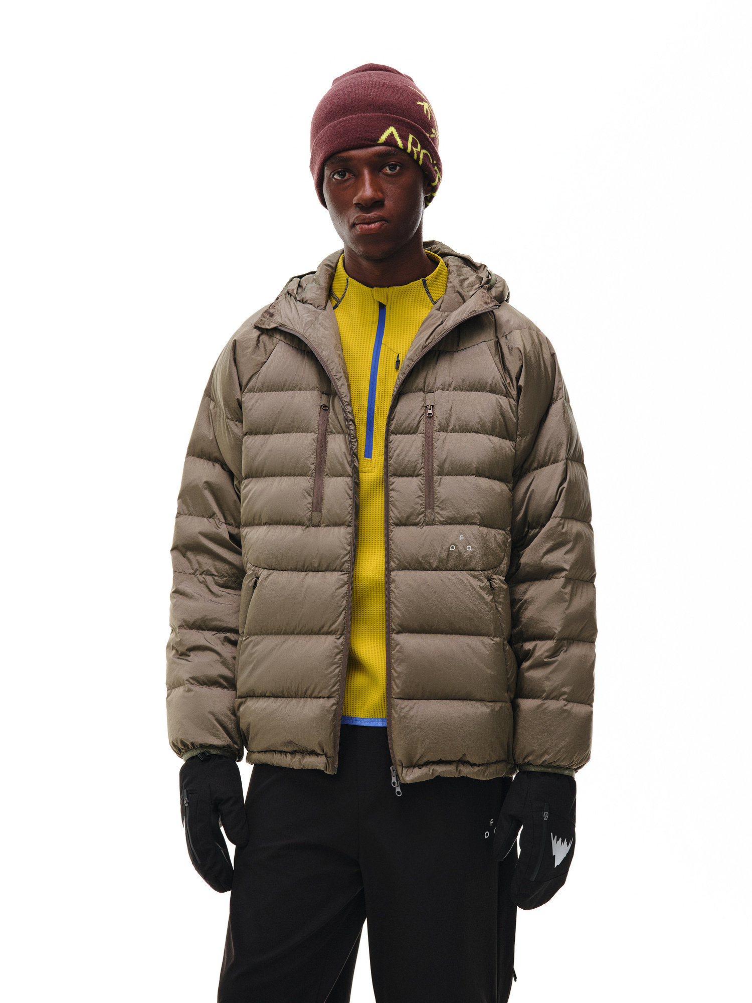 A person wears an EastLyst Dual-Zip Quilted Puffer Jacket over a yellow zip-up sweater, paired with black pants, black gloves, and a maroon beanie, standing against a plain white background.