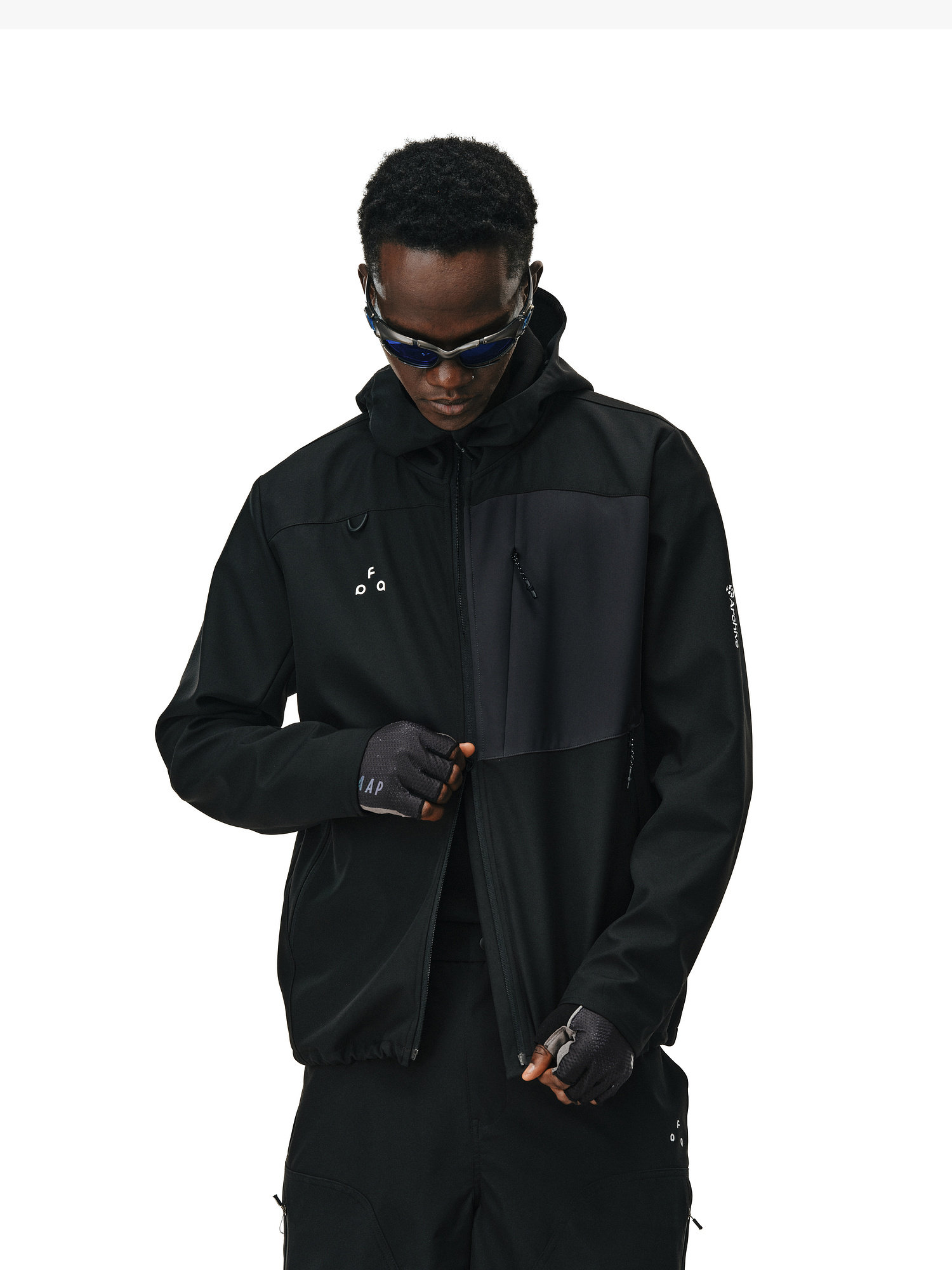 A person in EastLyst's Color-Block Water-Resistant Softshell Jacket, dark sunglasses, black pants, and fingerless gloves stands against a white background, looking down as they adjust their jacket.