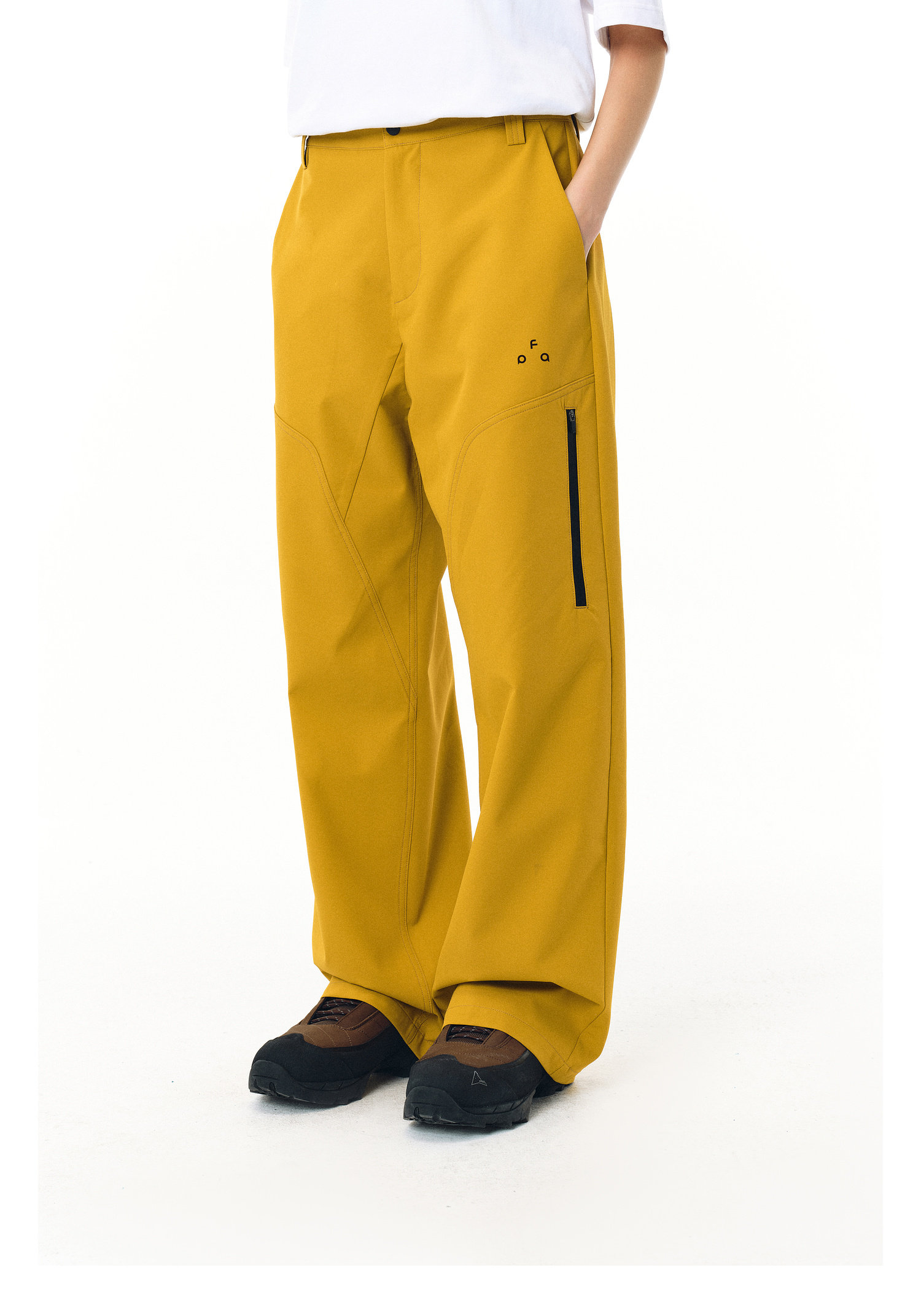 A person wears EastLyst Waterproof Windproof Softshell Pants in mustard yellow with wide legs, a side zipper, and small dot detail on the left thigh. They pair it with a white shirt and brown shoes, hands in pockets, against a white background.