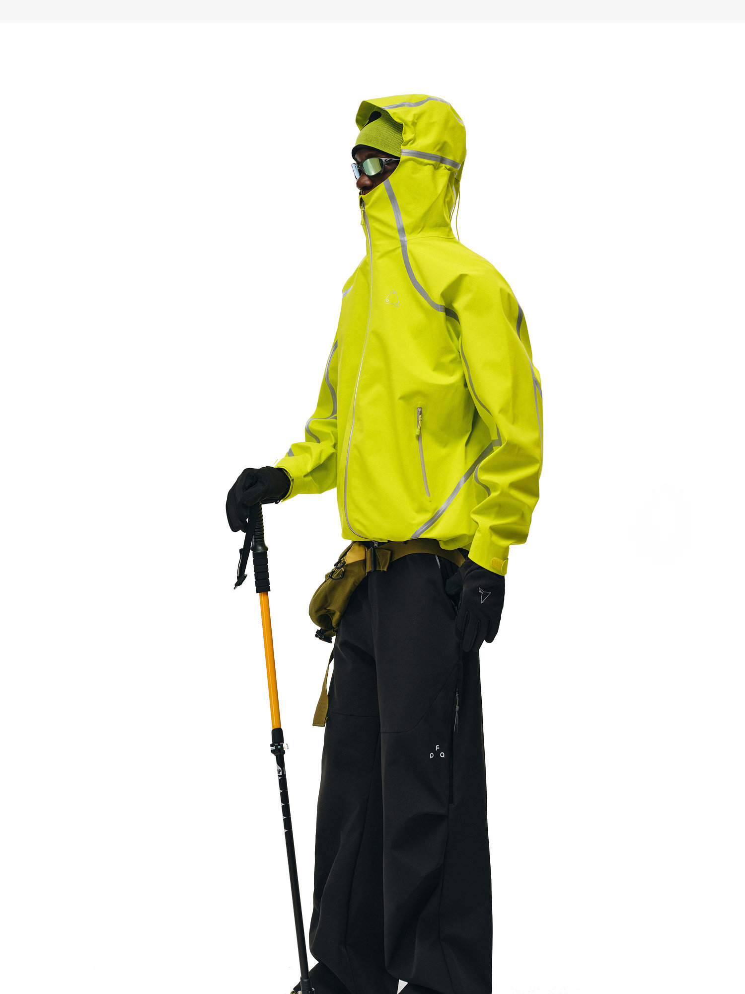 Wearing EastLyst Softshell Joggers, black gloves, sunglasses, and a bright yellow waterproof polyester hooded jacket, a person stands sideways with a hiking pole against a white background.