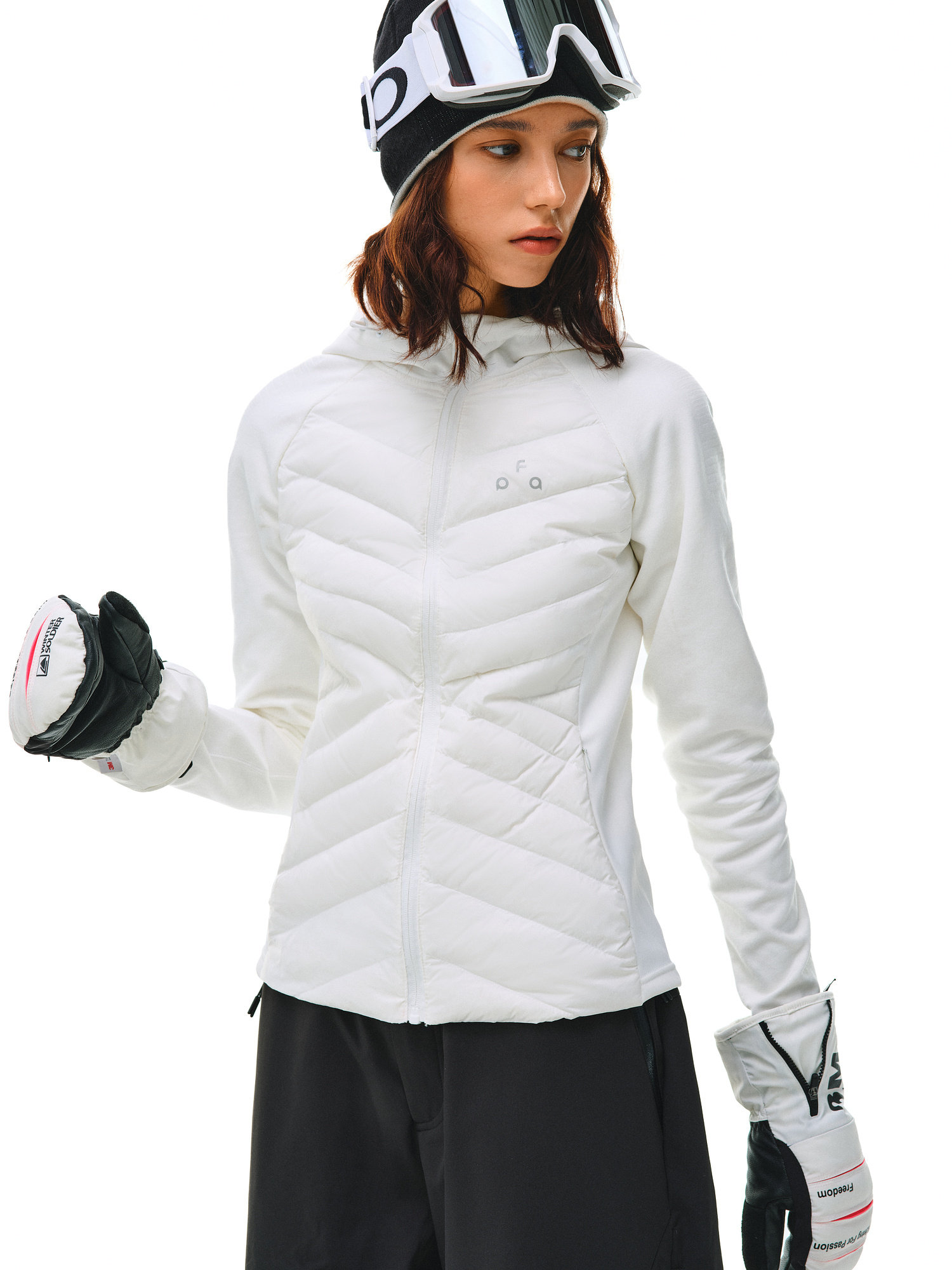 A woman in winter sportswear wears the EastLyst Quilted Hooded Down Jacket for lightweight warmth, paired with black snow pants, ski gloves, and a beanie with goggles. She faces sideways against a plain white background.