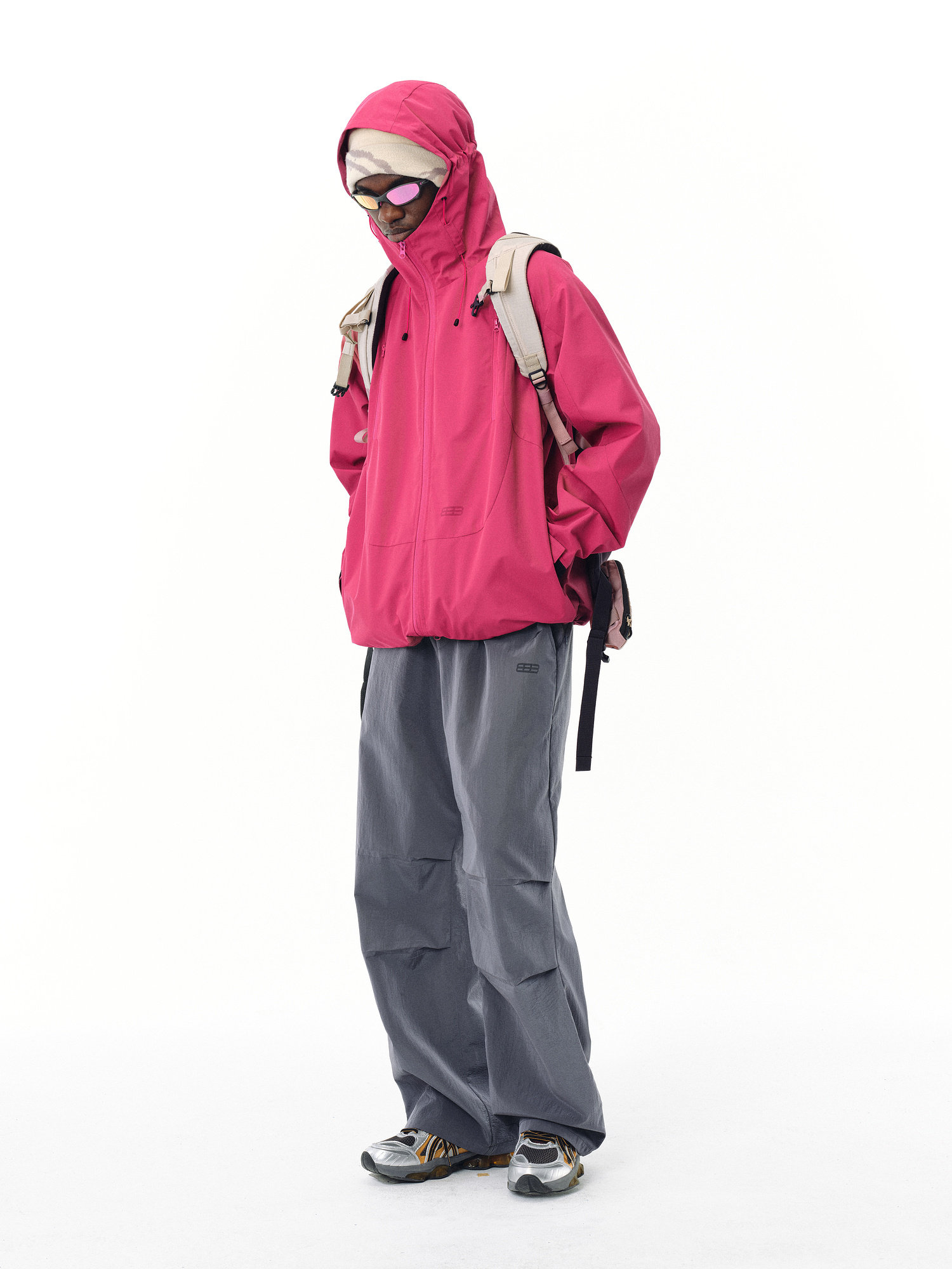 A person stands against a plain white background wearing the EastLyst Best All-Rounder Jacket in bright pink, paired with gray baggy pants, gray sneakers, sunglasses, a beige backpack, and a white cap.
