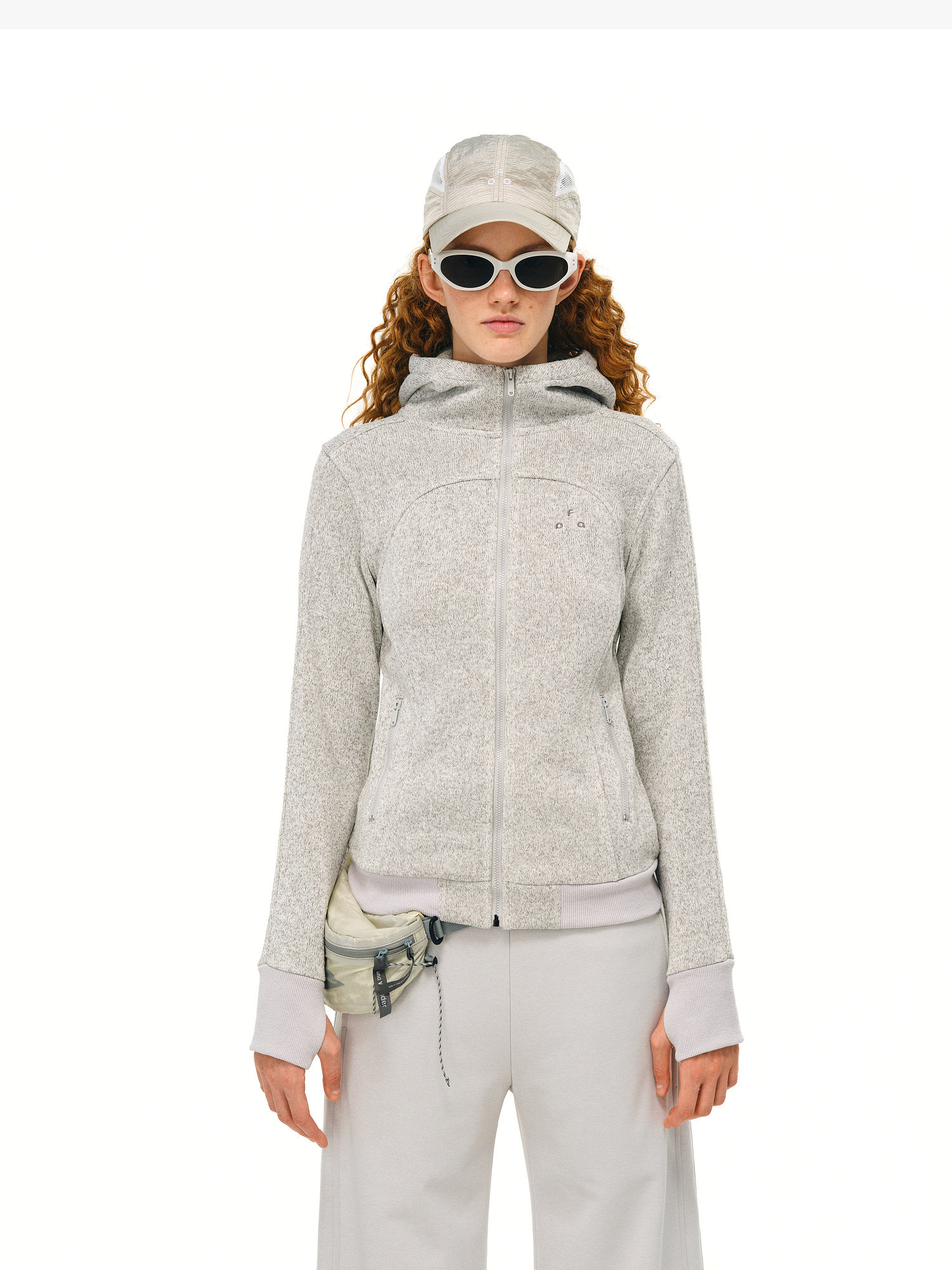 A woman with long curly hair wears the EastLyst Cropped Hooded Zip-Up Fleece Jacket in light gray and matching pants, accessorized with a tie-dye pouch, white oval sunglasses, and a gray cap. She stands against a plain white background.