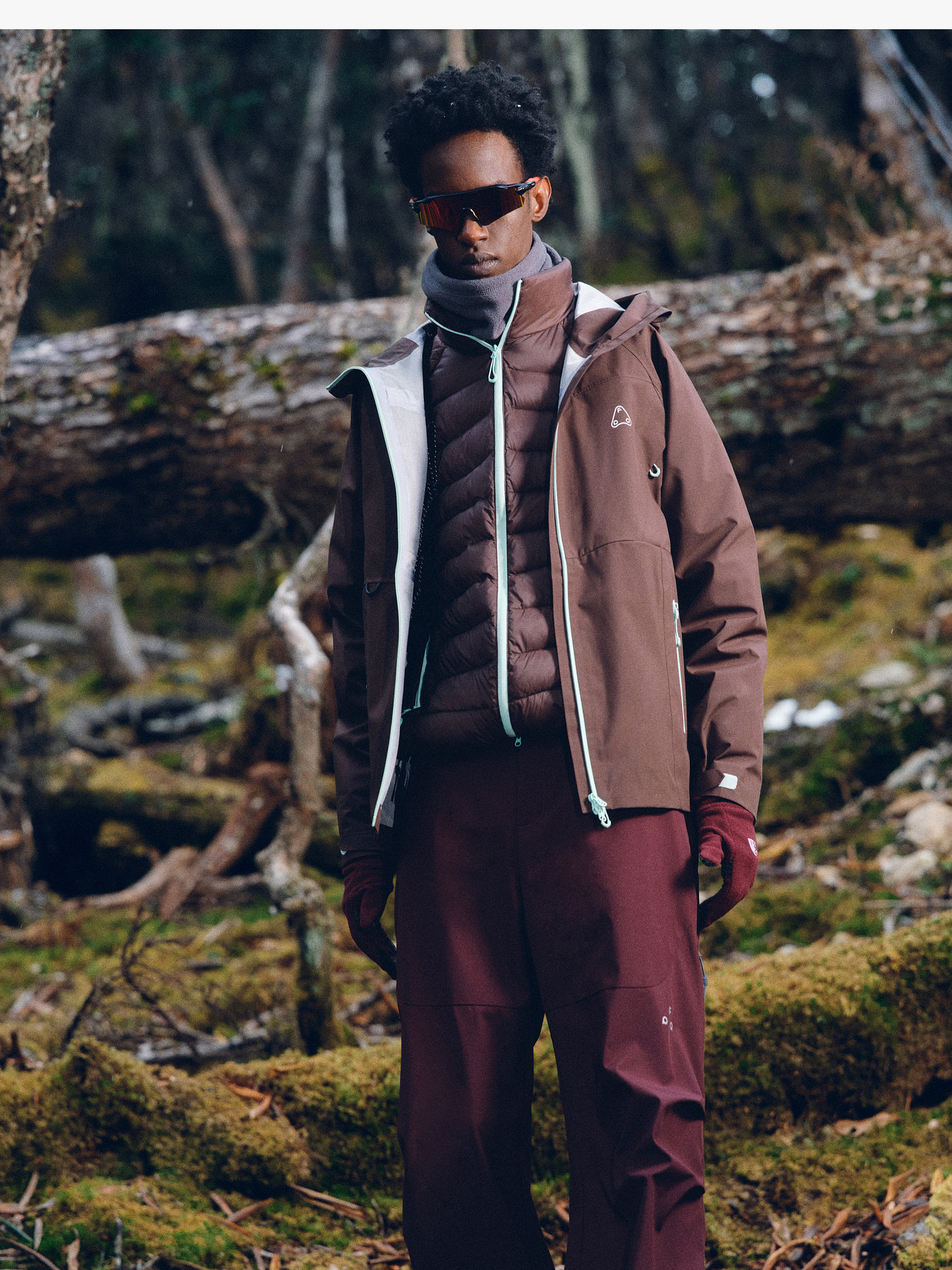 A person stands in a forest, wearing the EastLyst 3-in-1 Color-Block Storm Jacket, maroon pants, gloves, sunglasses, and a scarf, with mossy ground and fallen trees in the background.