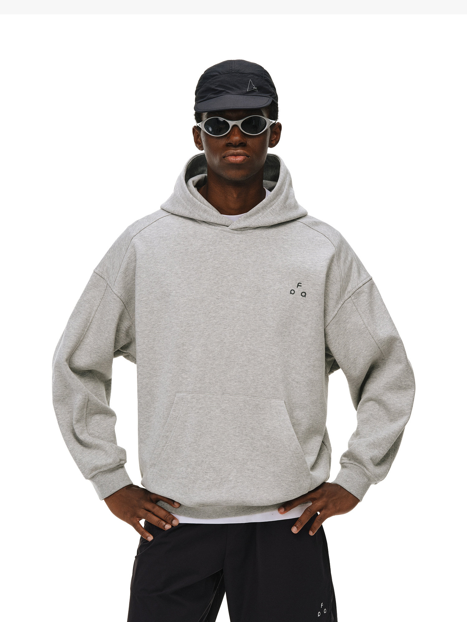 A person in an EastLyst Outdoor Print Hoodie, paired with a black cap and dark sunglasses, stands hands on hips against a plain white background.