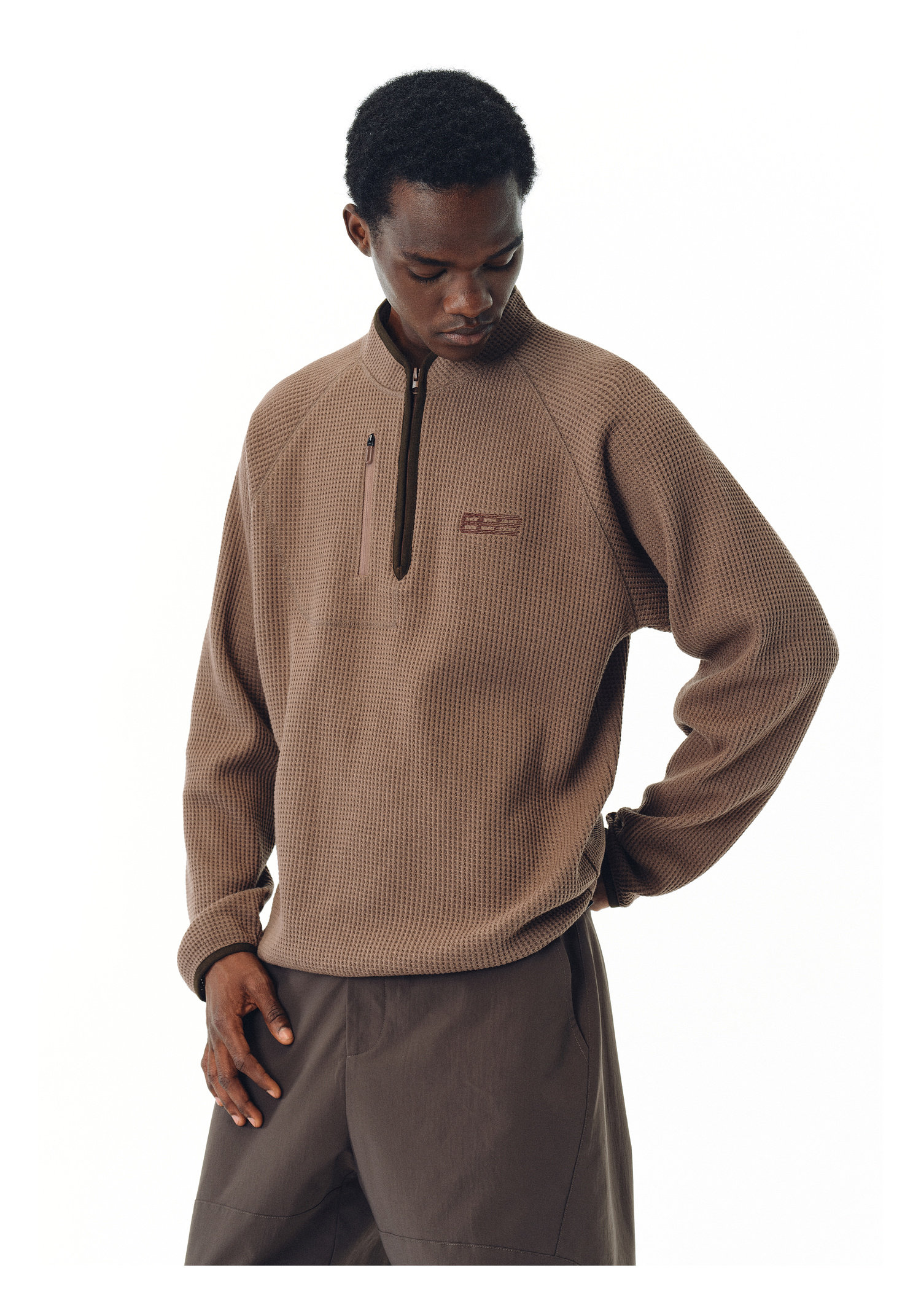 A man wearing the EastLyst Heavyweight Magnetic Waffle Knit Pullover Hoodie and dark brown pants stands against a plain white background, looking down with one hand on his hip.