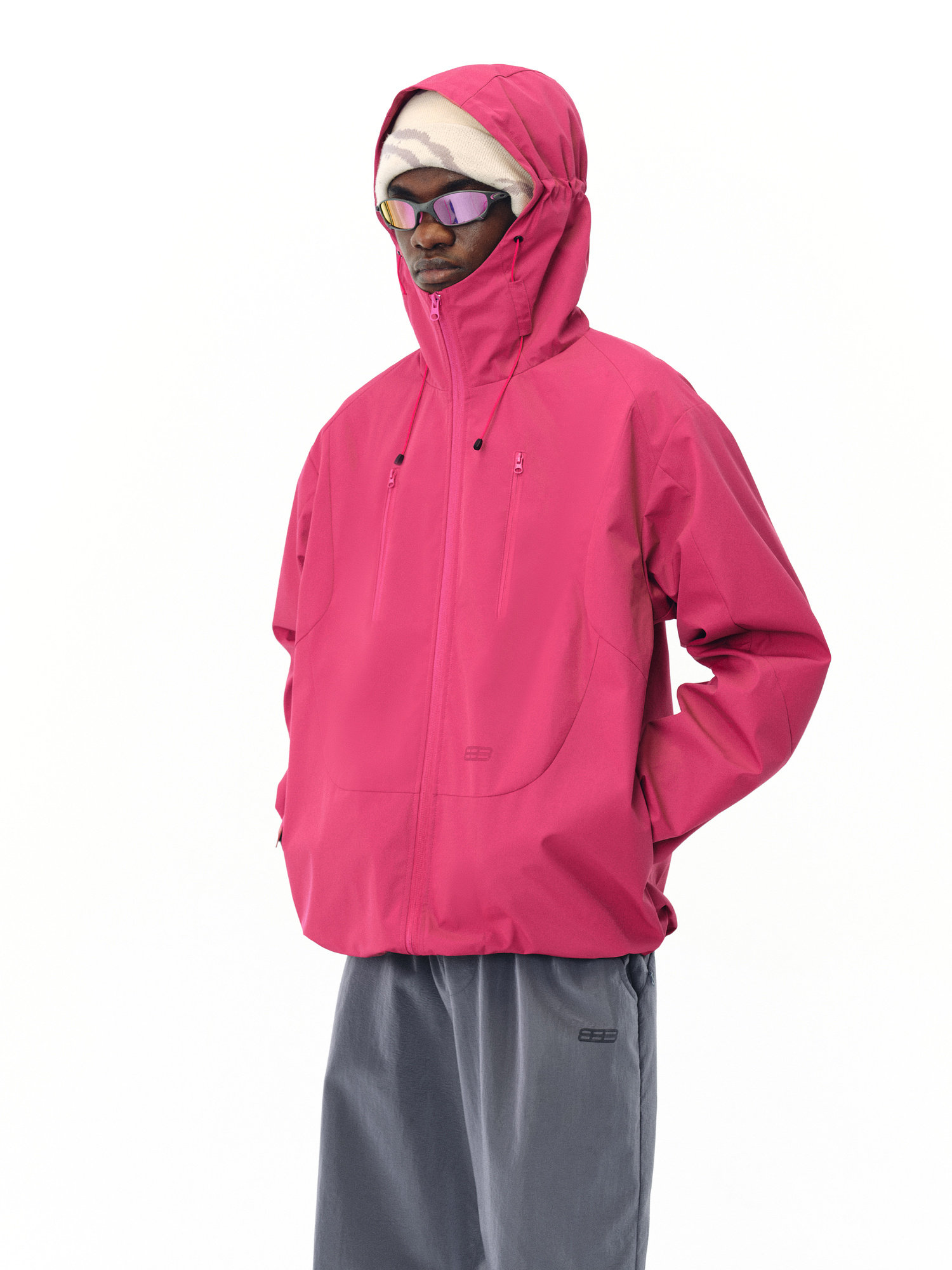 Against a plain white background, a person in loose gray pants wears the EastLyst Best All-Rounder Jacket in bright pink, paired with purple sunglasses and a cream beanie, standing with hands in jacket pockets.