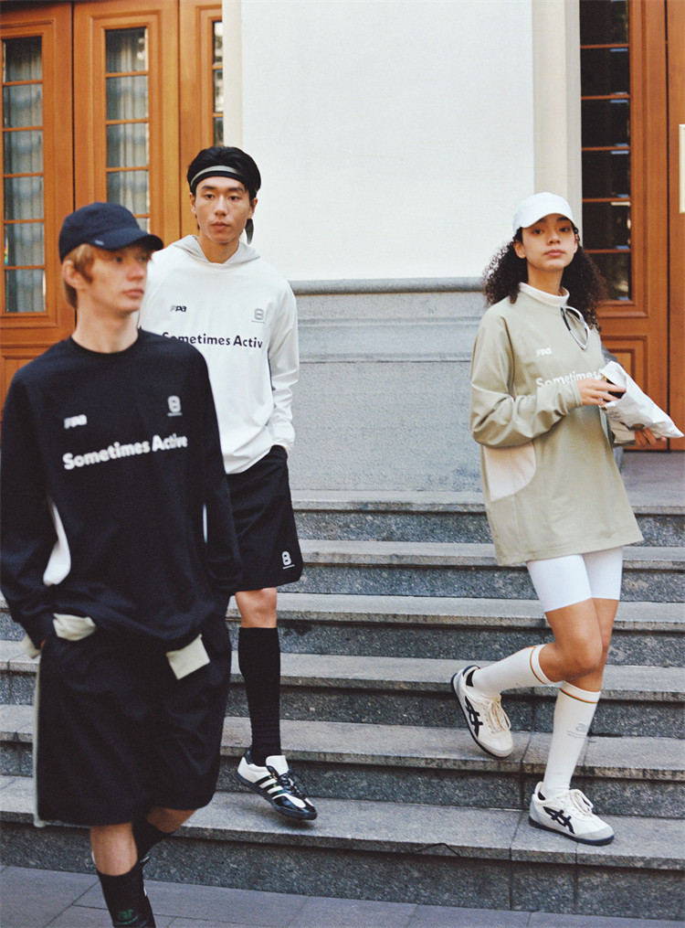 Three young adults in sporty, casual outfits stand and walk near stone steps outside a building. They wear caps and oversized EastLyst Quick-Dry Color Block Long Sleeve Tees with "Sometimes Active" printed on them.