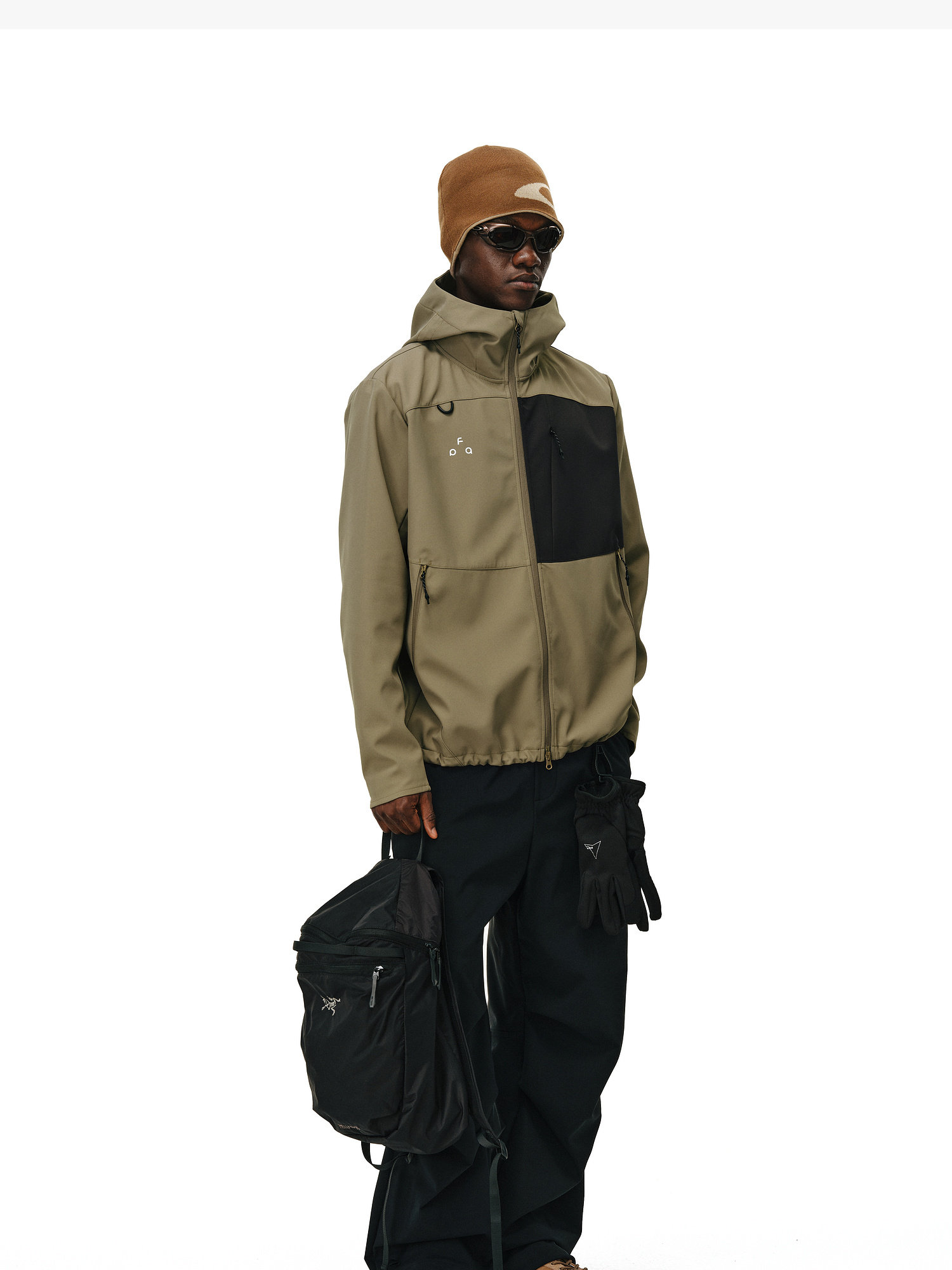 A person in sunglasses, a tan beanie, EastLyst Color-Block Water-Resistant Softshell Jacket, black pants, and dark gloves holds a black backpack against a plain white background.