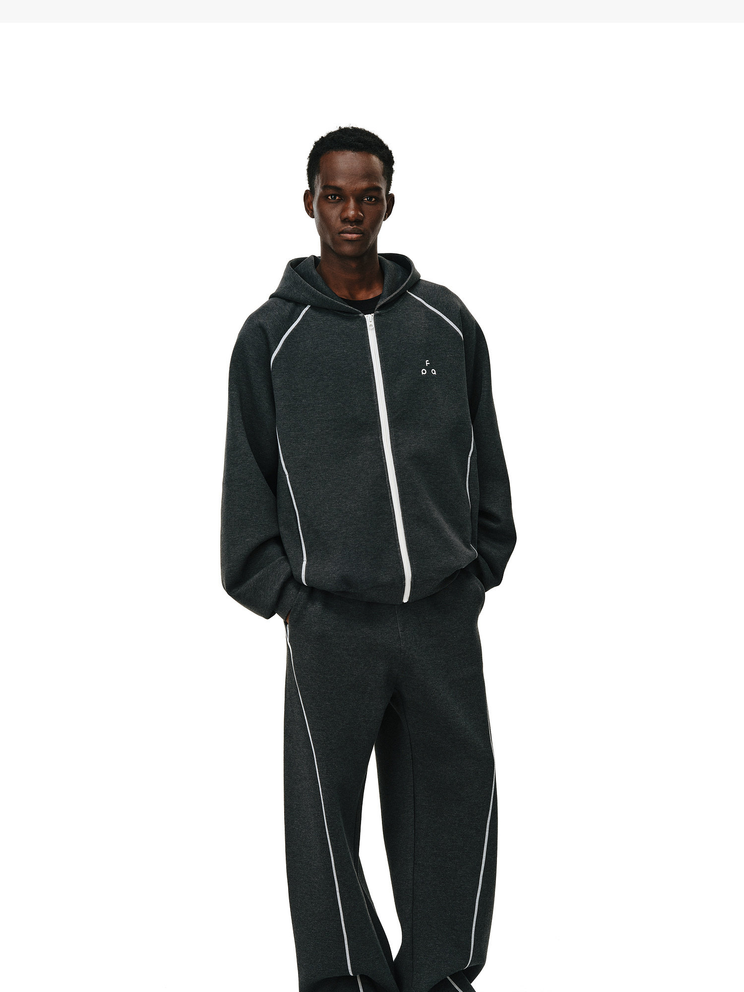 A person wears the EastLyst Constant Warmth Zip Hoodie, a dark gray tracksuit with white piping, standing against a plain white background. The high-end fabric brings luxury and comfort to this stylish daily essential.