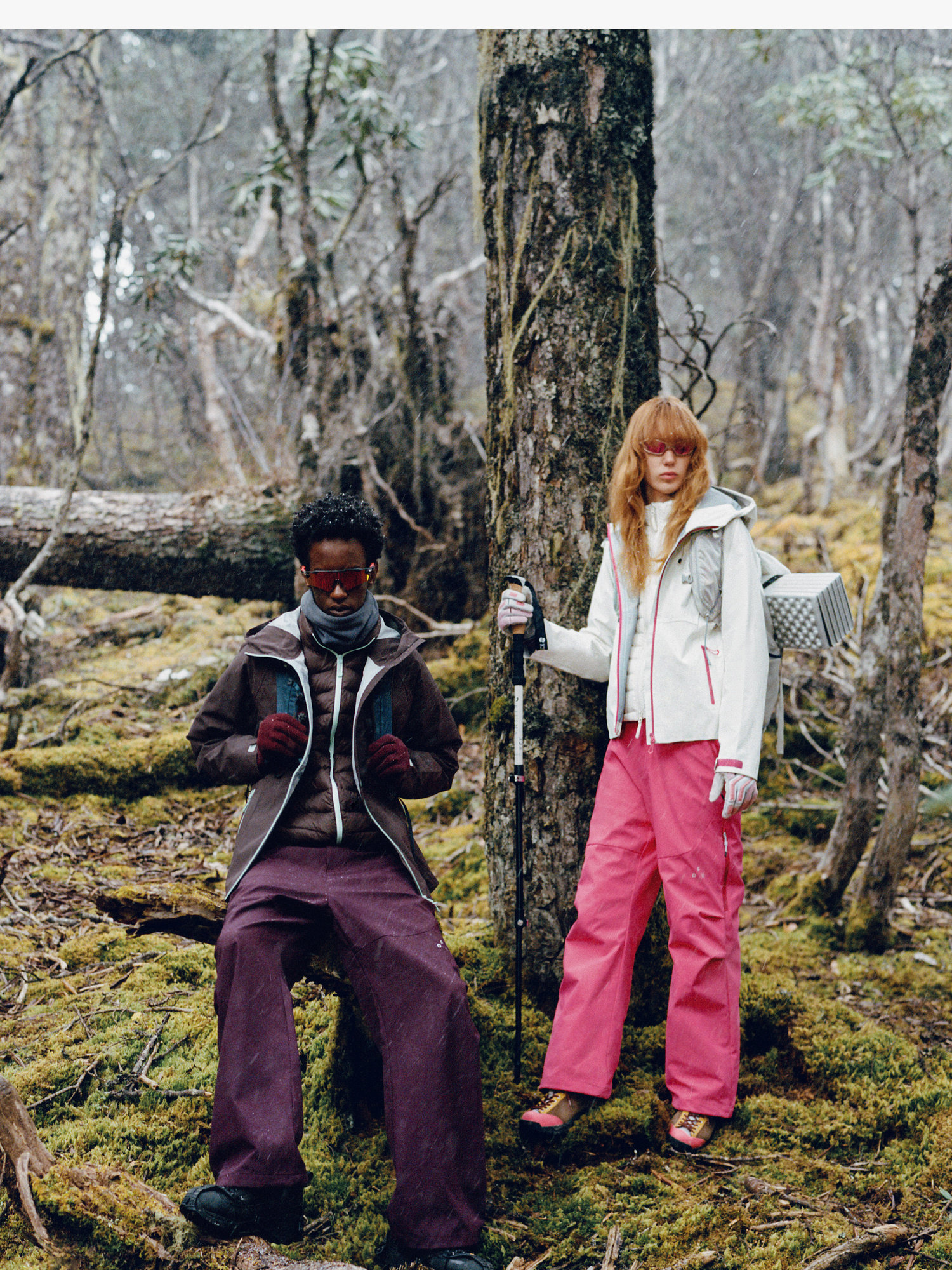Two people in a mossy forest wear EastLyst 3-in-1 Color-Block Storm Jackets. One sits on a log, the other stands with a hiking pole—both ready for any adventure amid trees and greenery.