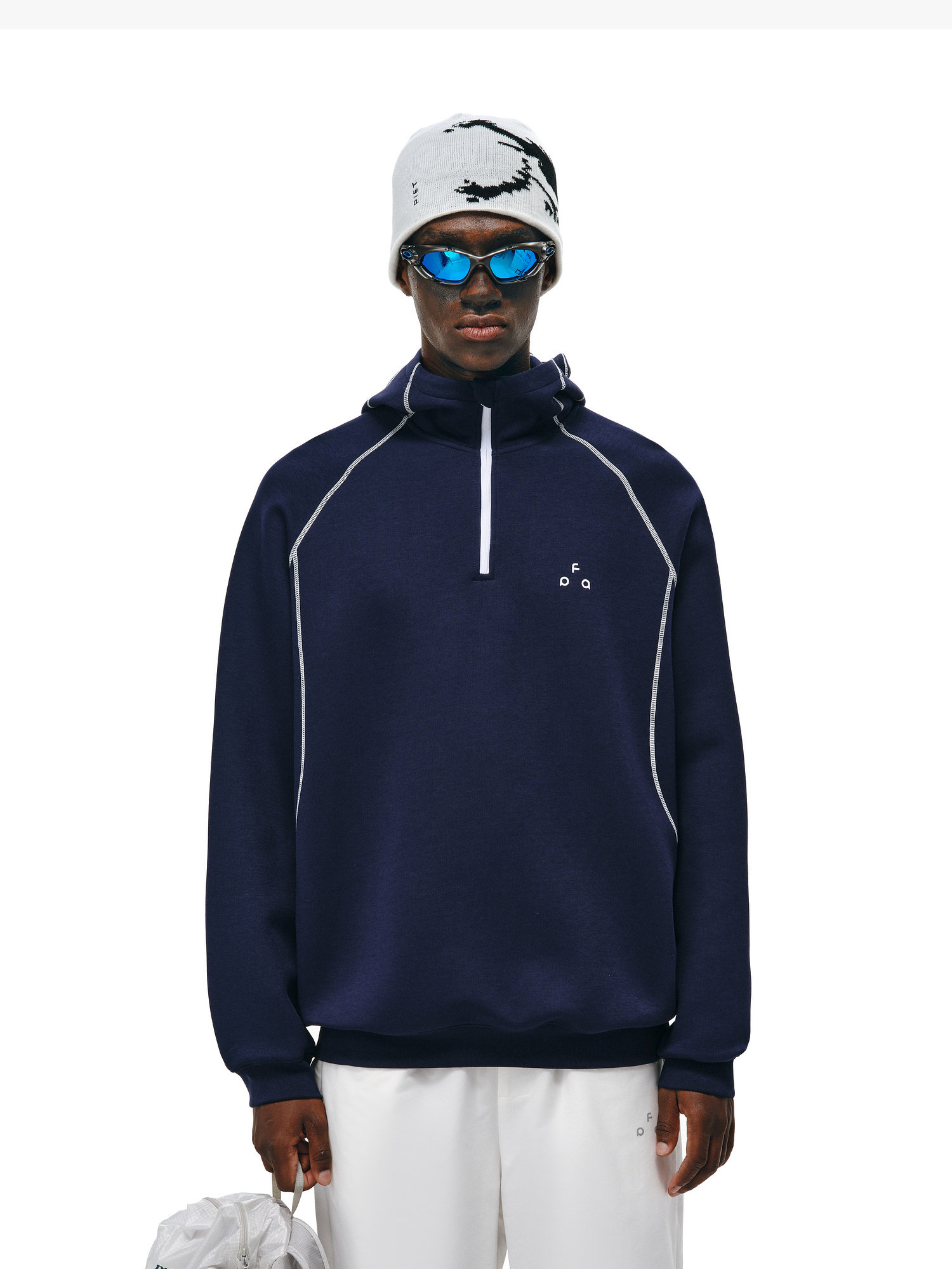 A person wearing the EastLyst Retro-Core Half-Zip Hoodie in navy blue with white accents, paired with white pants, blue reflective sunglasses, and a white beanie with black pattern, stands against a plain white background.
