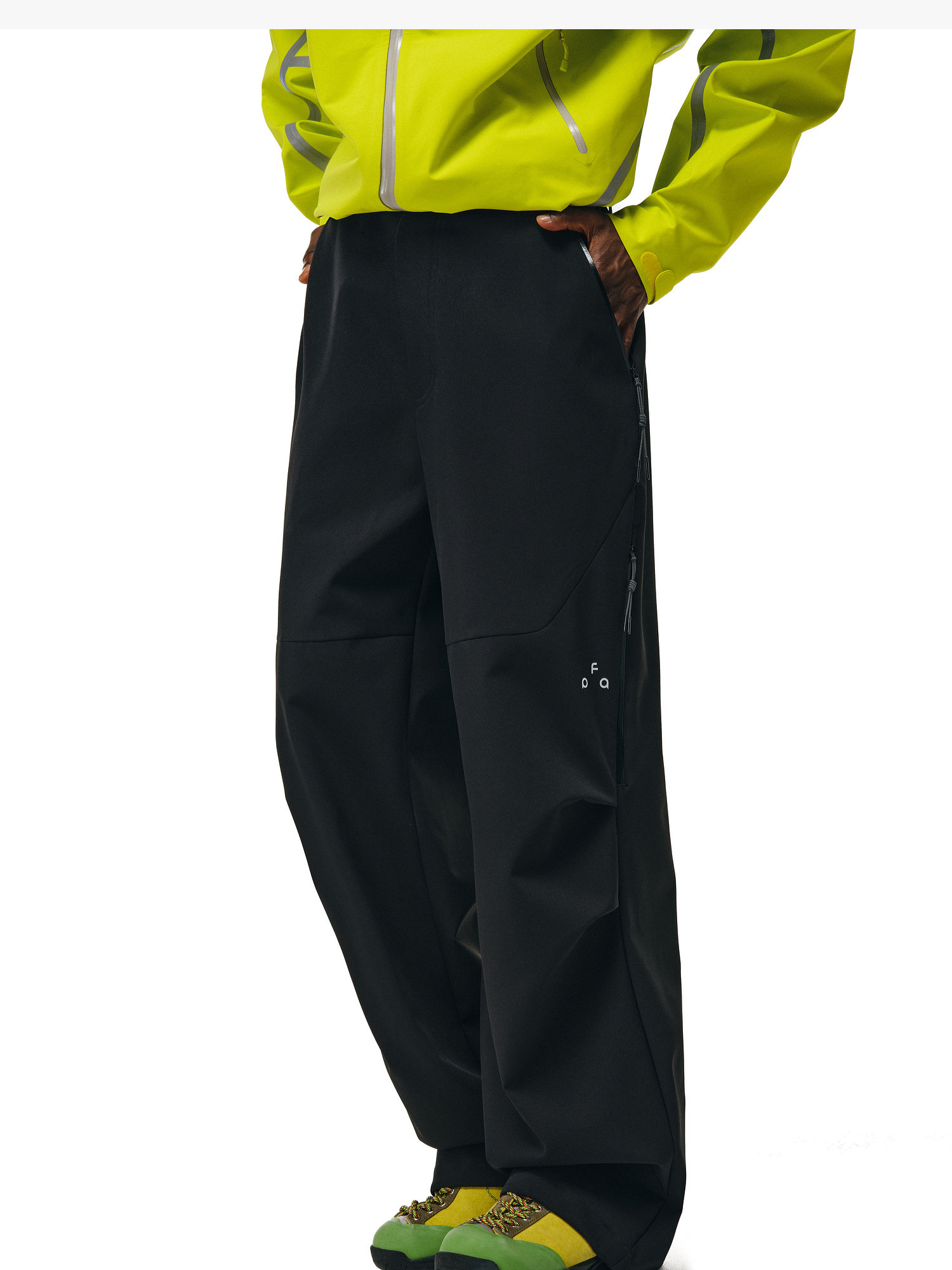 A person stands with hands in pockets against a white background, wearing Softshell Joggers by EastLyst, a bright yellow waterproof polyester jacket, and green-and-yellow sandals.