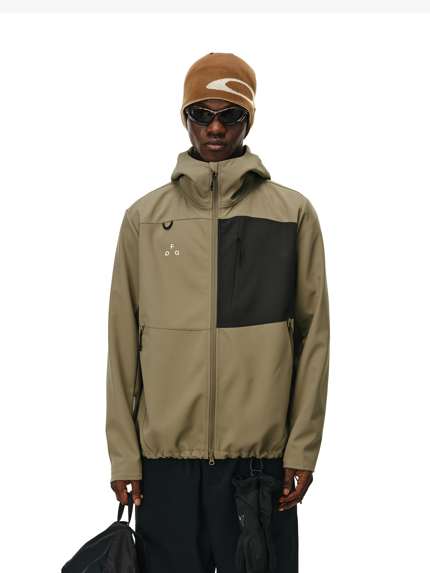 A person wears the EastLyst Color-Block Water-Resistant Softshell Jacket, a tan beanie with a white design, black sunglasses, and dark pants, standing against a white background and holding a black bag in their left hand.