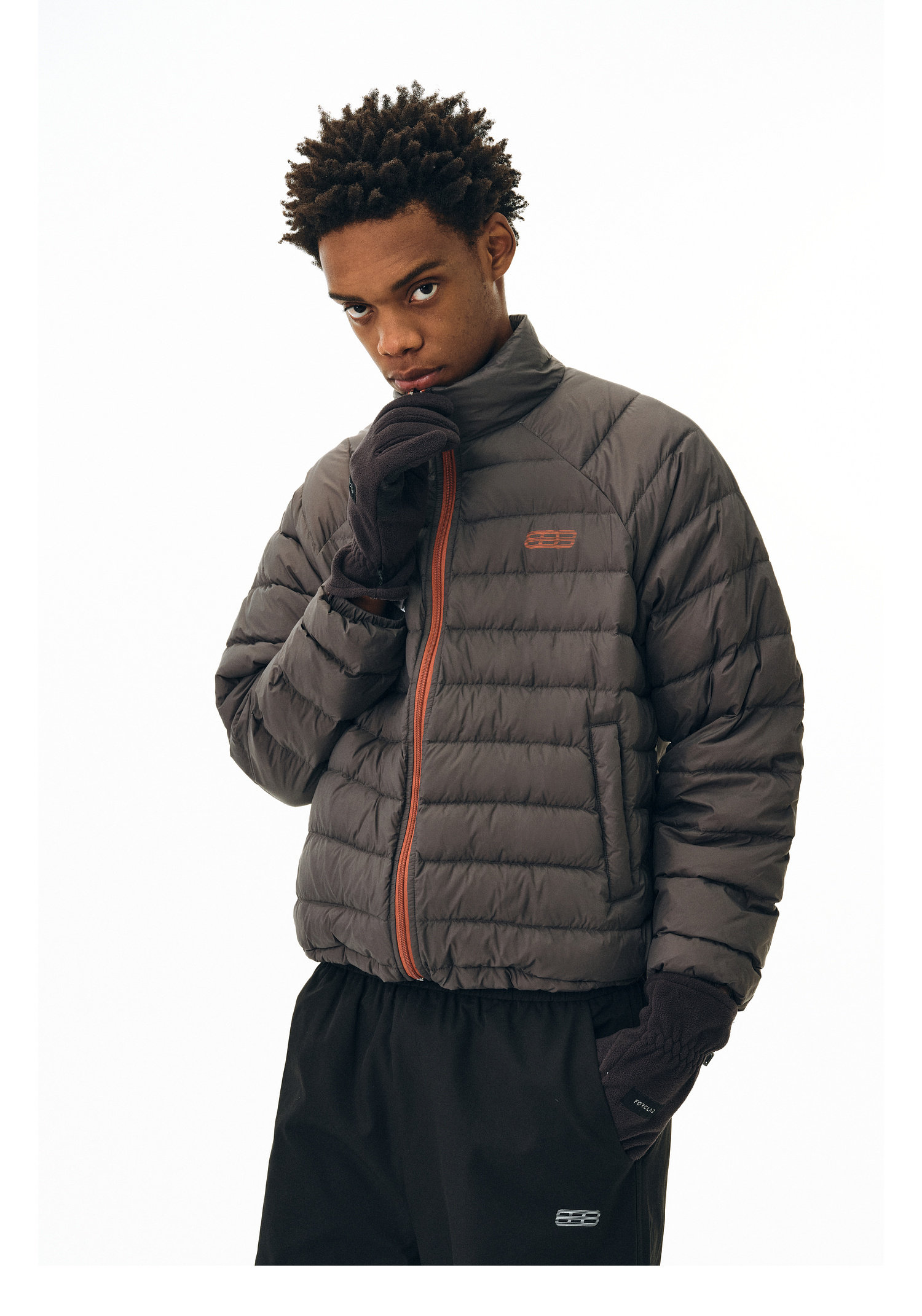 A person wears the EastLyst Versatile Explorer 3-in-1 Jacket in dark color with an orange zipper and matching gloves, standing against a plain white background while holding the collar and looking at the camera.