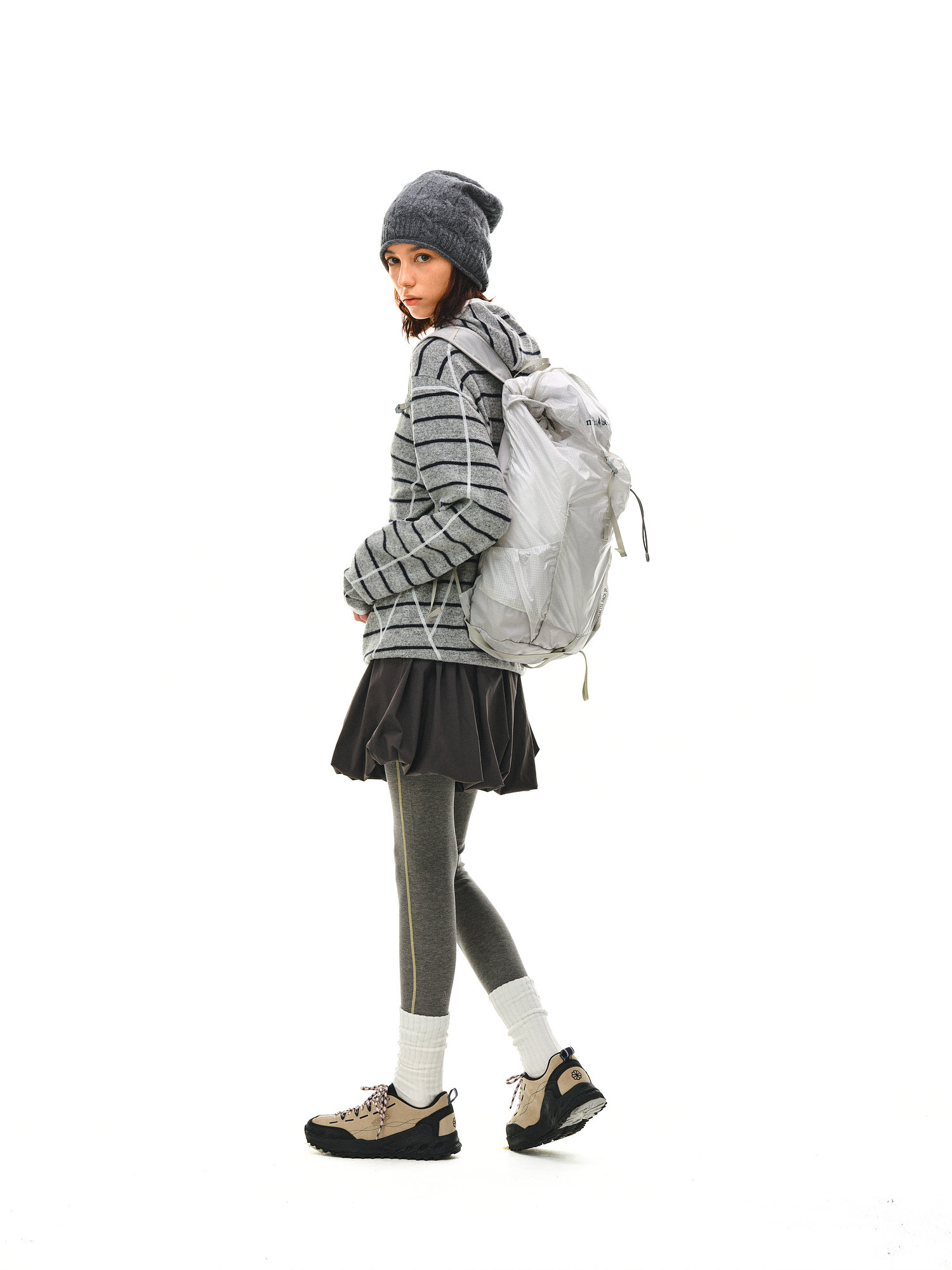 A woman in the EastLyst Textured Stripe Knit Hoodie stands sideways against a white background, pairing it with a gray beanie, backpack, gray leggings, skirt, white socks, and hiking boots for an effortlessly casual look.