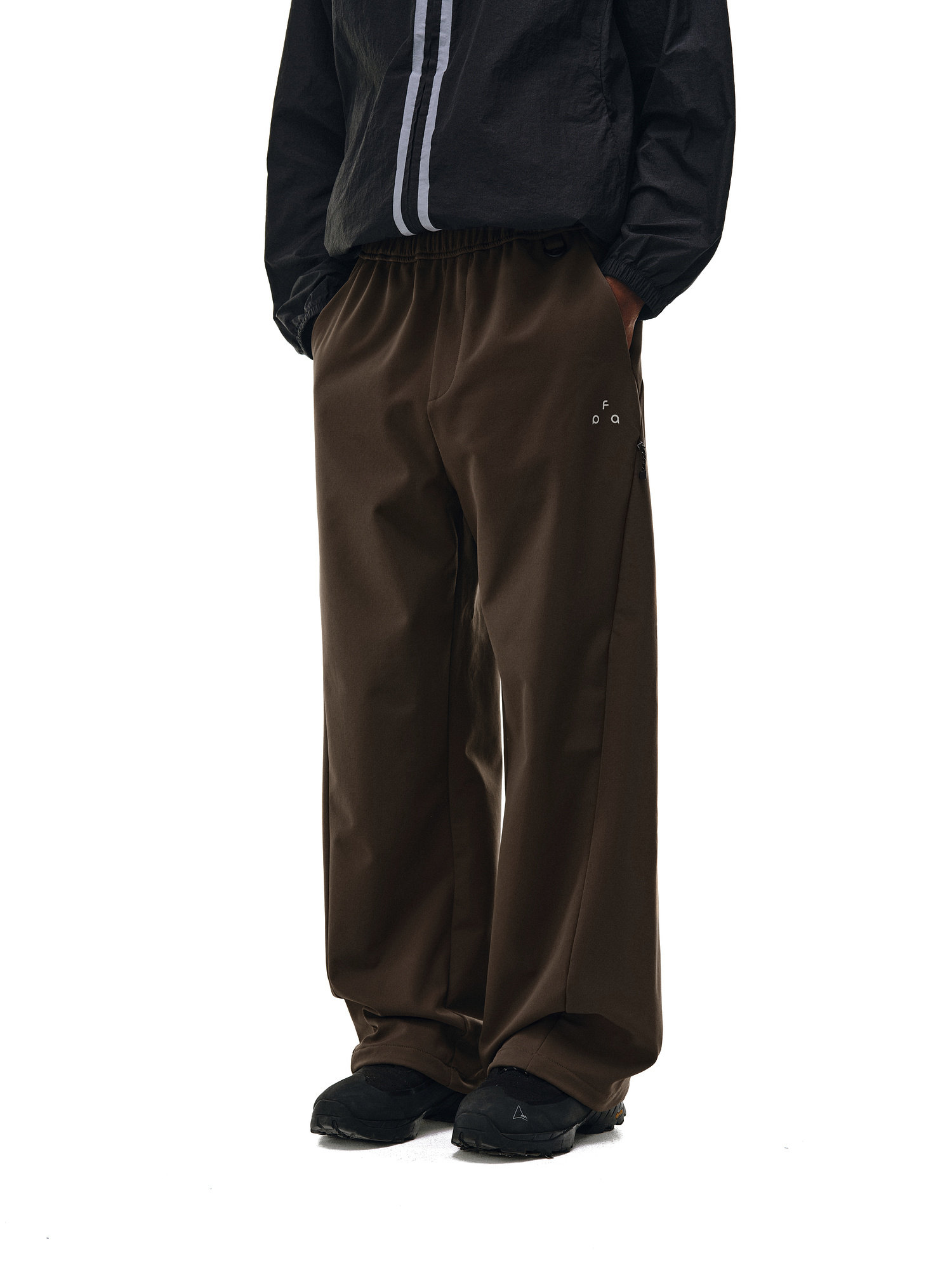 A person wears EastLyst Urban Outdoor Softshell Pants in relaxed fit, dark brown, paired with a black jacket featuring a vertical gray stripe and black shoes, posing against a plain white background.