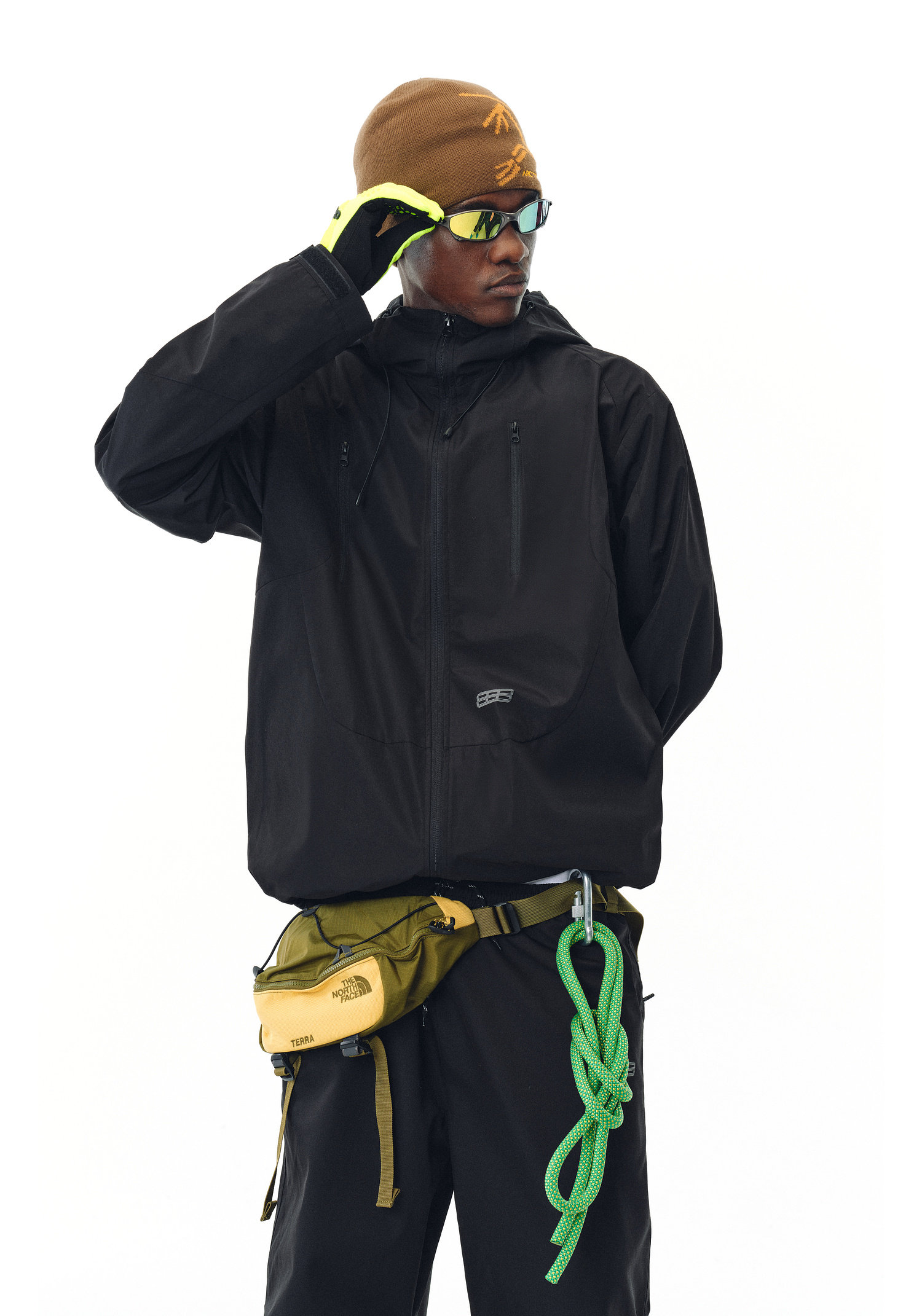 A person in an EastLyst Best All-Rounder Jacket, brown beanie, black pants, and neon green gloves stands against a white background, sporting sunglasses, a yellow waist bag, and a green climbing rope clipped to their belt.