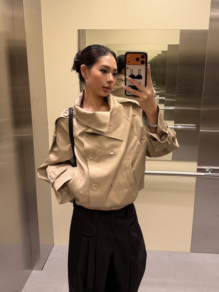 Woman wearing a beige cropped trench jacket with a black pleated skirt in an elevator mirror selfie