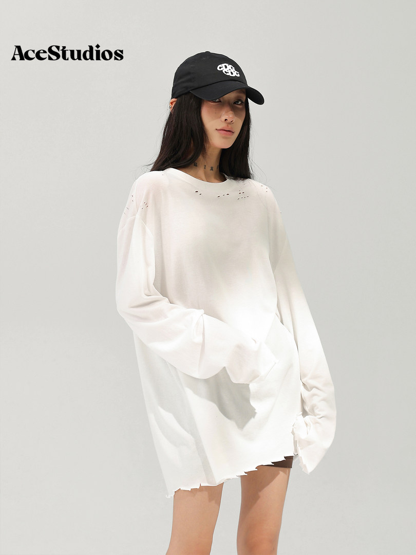 Model wearing an oversized distressed white long-sleeve top with a black baseball cap.