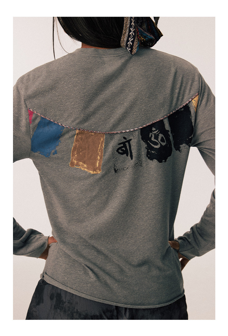 A person wearing the EastLyst Vintage vintage T-shirt, featuring a gray long-sleeve design with vibrant prayer flag print and black symbols on the upper back, stands hands on hips with their back to the camera.