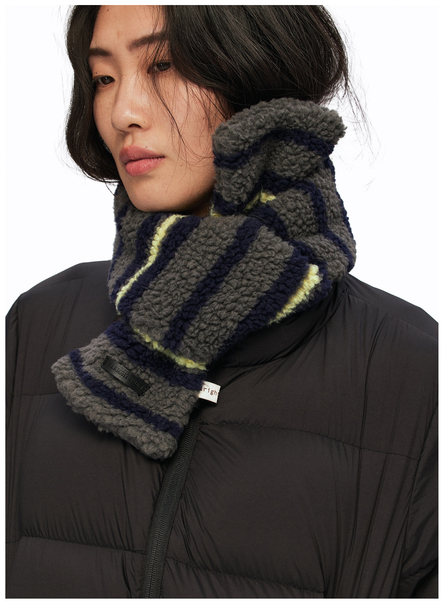A woman with long dark hair and a neutral expression wears a black puffer jacket and the EastLyst Striped Fleece Scarf, featuring gray stripes with navy and yellow accents, wrapped snugly around her neck for a cozy look.