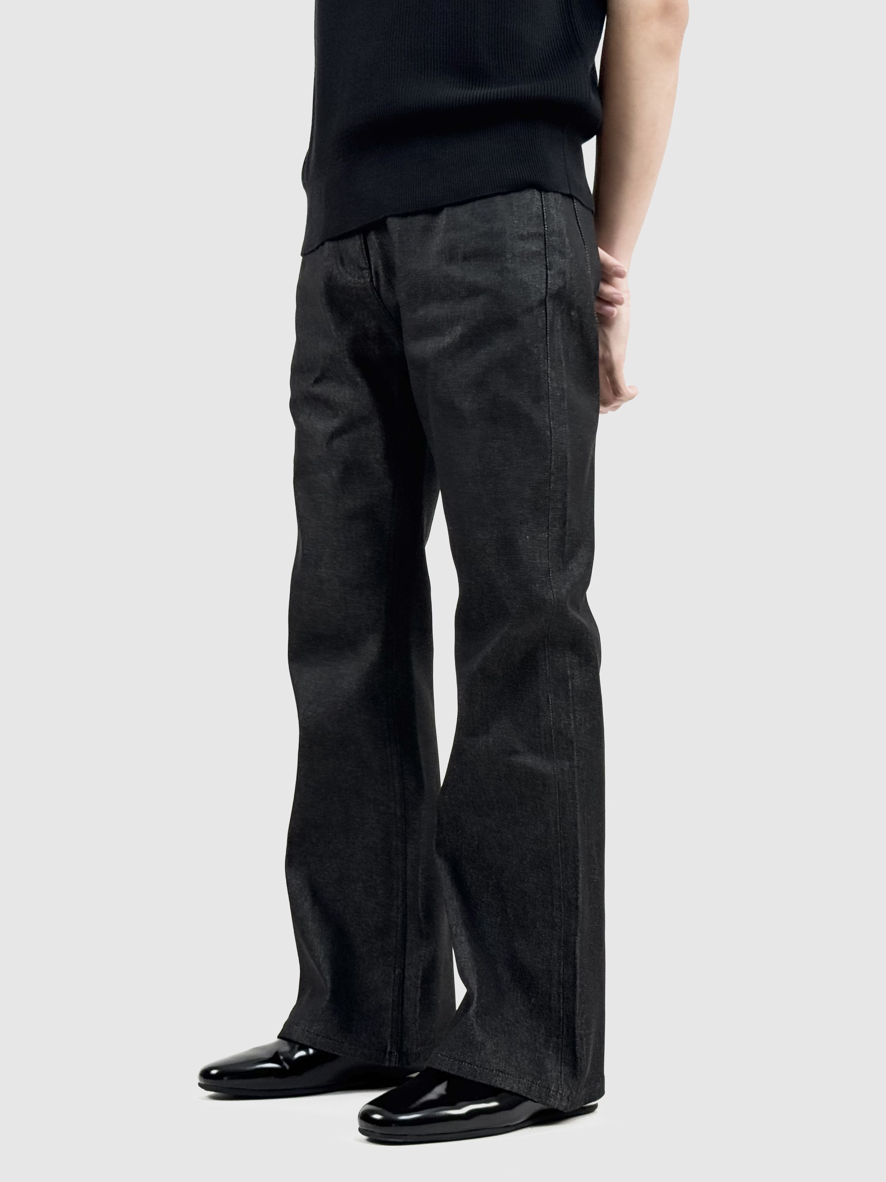 A person wears EastLyst Colorfast Black Flared Jeans and black shiny shoes, hands behind their back, shown from the chest down against a plain light gray background.