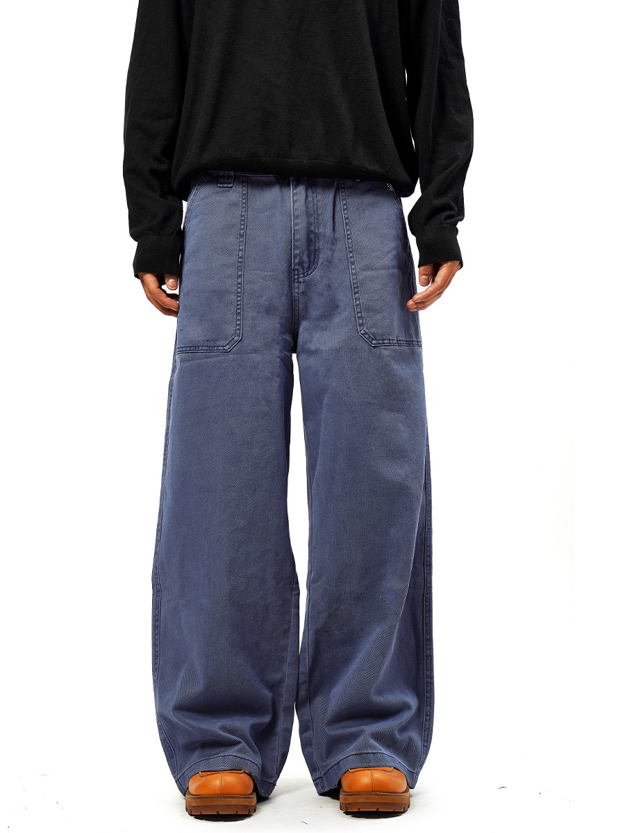 A person wearing a black long-sleeve shirt, EastLyst French Workwear Pants—loose-fitting blue wide-leg jeans with a distressed finish—and brown shoes, stands against a plain white background.