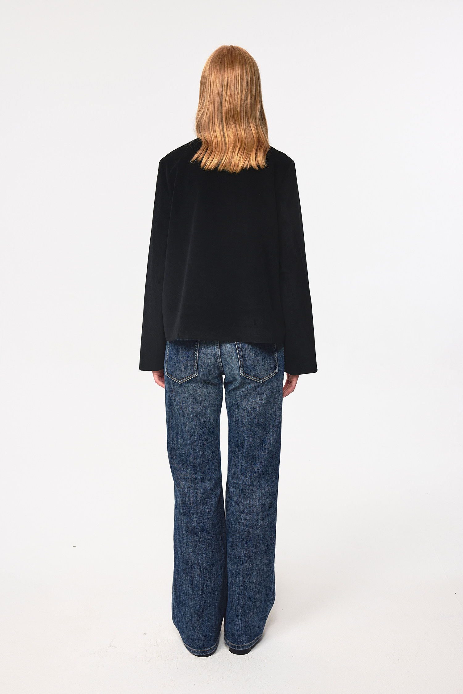 A person with shoulder-length blonde hair faces away, wearing a black jacket and EastLyst Slub Textured Casual Pants in vintage indigo wash against a plain white background.