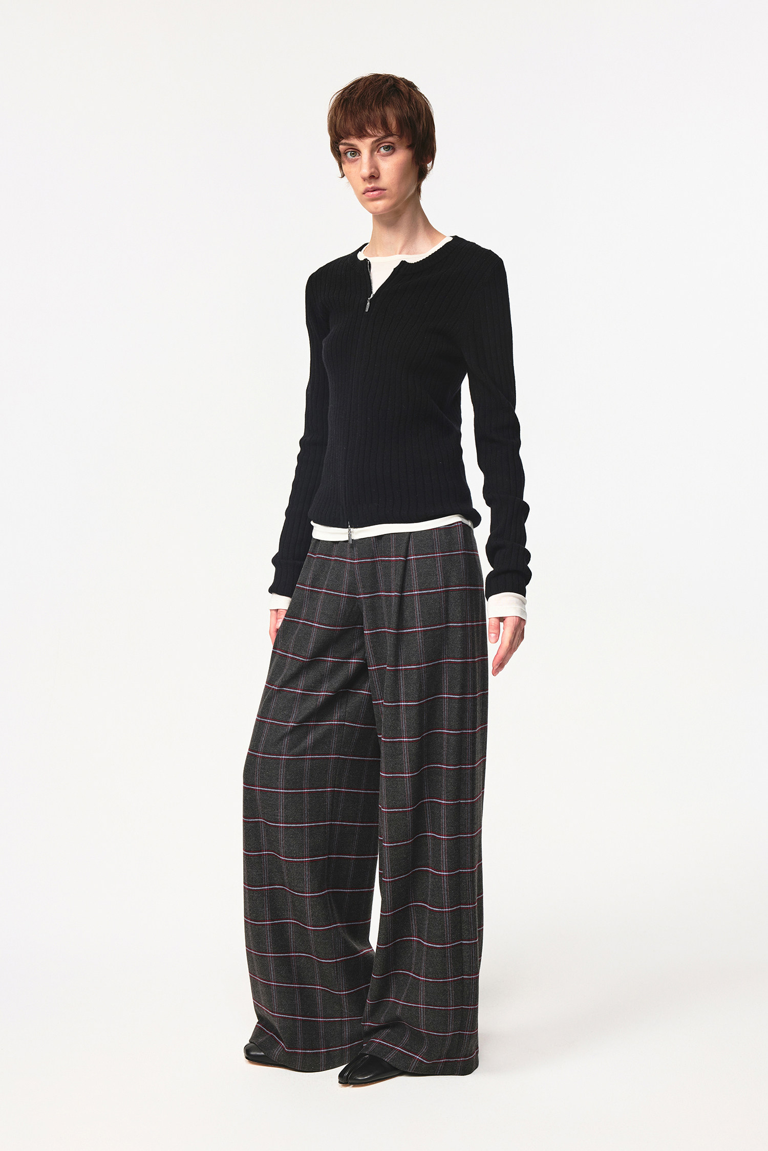 A person with short brown hair wears the EastLyst Merino Wool Cardigan, featuring a ribbed texture and white trim, paired with loose plaid trousers. They stand against a plain white background, looking directly at the camera.