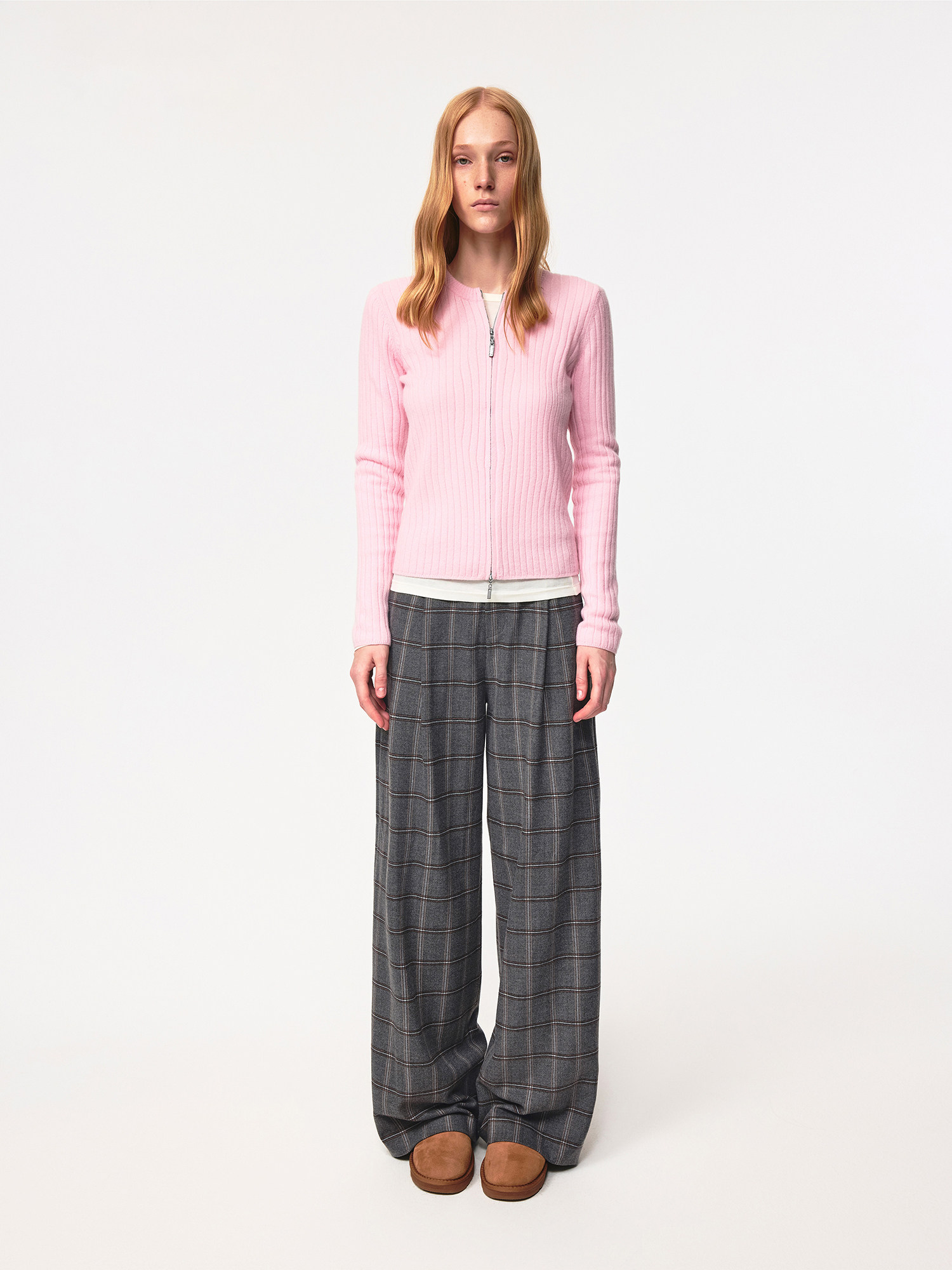 A woman with long blonde hair stands against a plain backdrop, wearing the EastLyst Merino Wool Cardigan in light pink, paired with gray wide-leg plaid pants and brown slip-on shoes. She faces forward with a neutral expression.