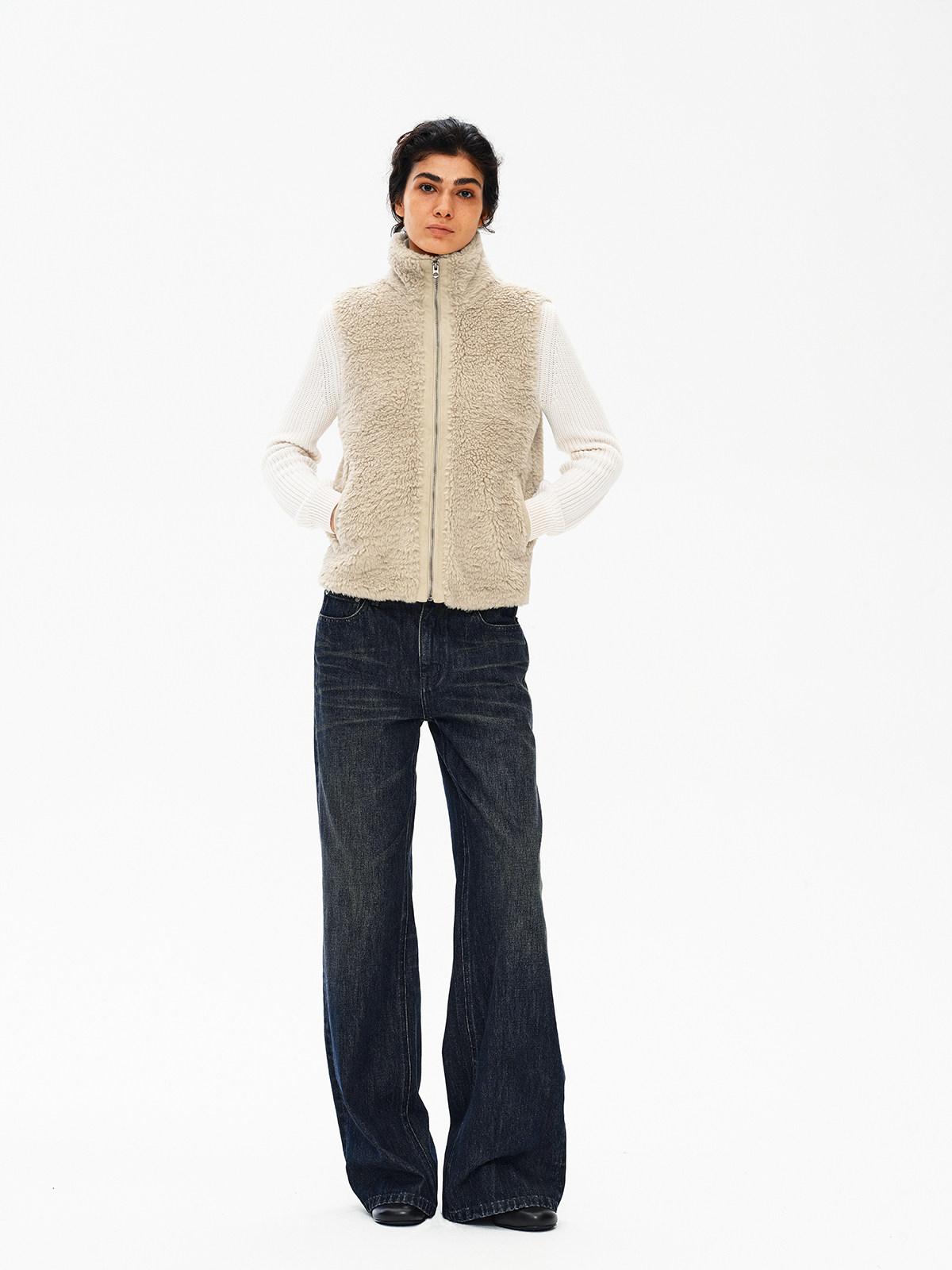一个人站在白色背景前，双手插在口袋里，穿着 EastLyst 米色高领羊羔绒背心，内搭白色毛衣和深色阔腿牛仔裤。