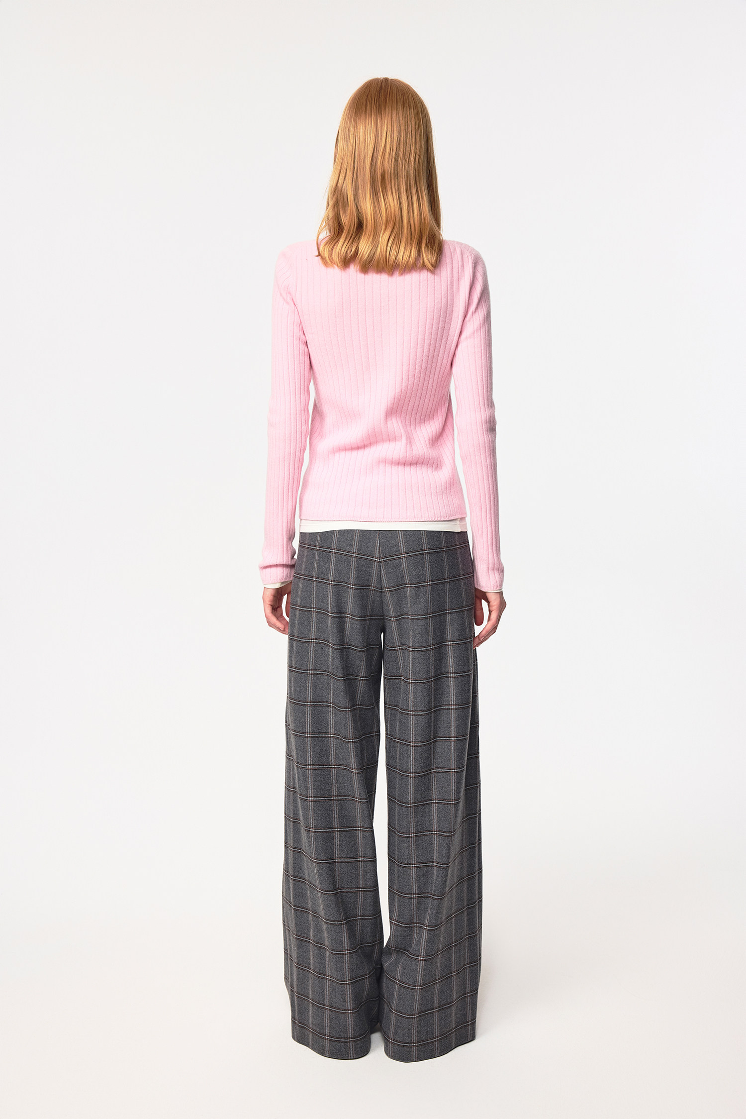 A person with shoulder-length blonde hair stands facing away, wearing the EastLyst Merino Wool Cardigan in light pink and wide-leg gray plaid pants, set against a plain white background.