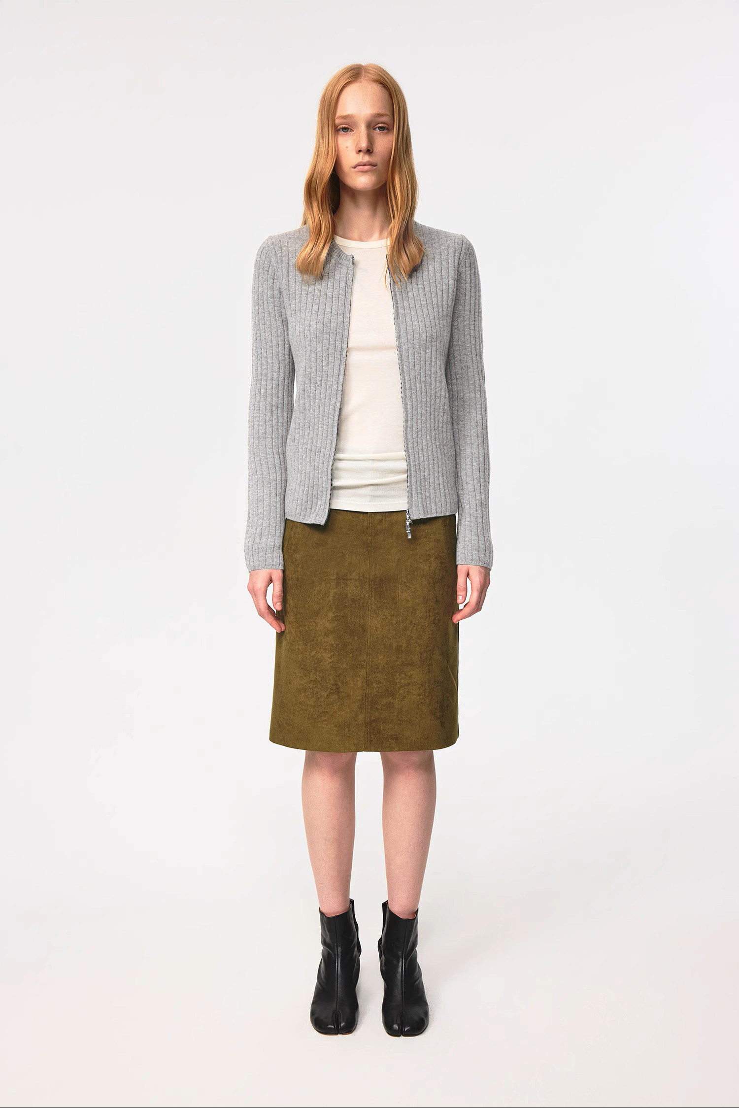 A woman with long blond hair stands facing forward, wearing the EastLyst Merino Wool Cardigan in gray over a white top, paired with an olive green knee-length skirt and black ankle boots against a plain light background.