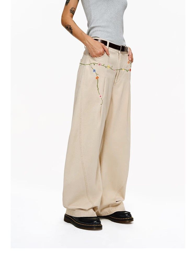 Only the lower half of a person is visible, wearing EastLyst Floral Embroidery Cargo Pants with colorful floral designs, paired with a black belt, black shoes, and a grey sleeveless top against a plain white background.