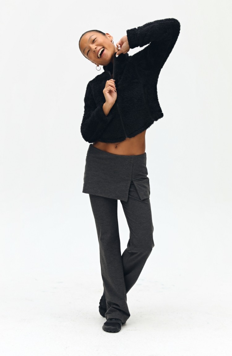 Wearing an EastLyst Pink Short Faux Shearling Jacket and a dark gray skirt over matching pants, a person smiles with eyes closed and arms raised near their face, posing cheerfully against a plain white background.