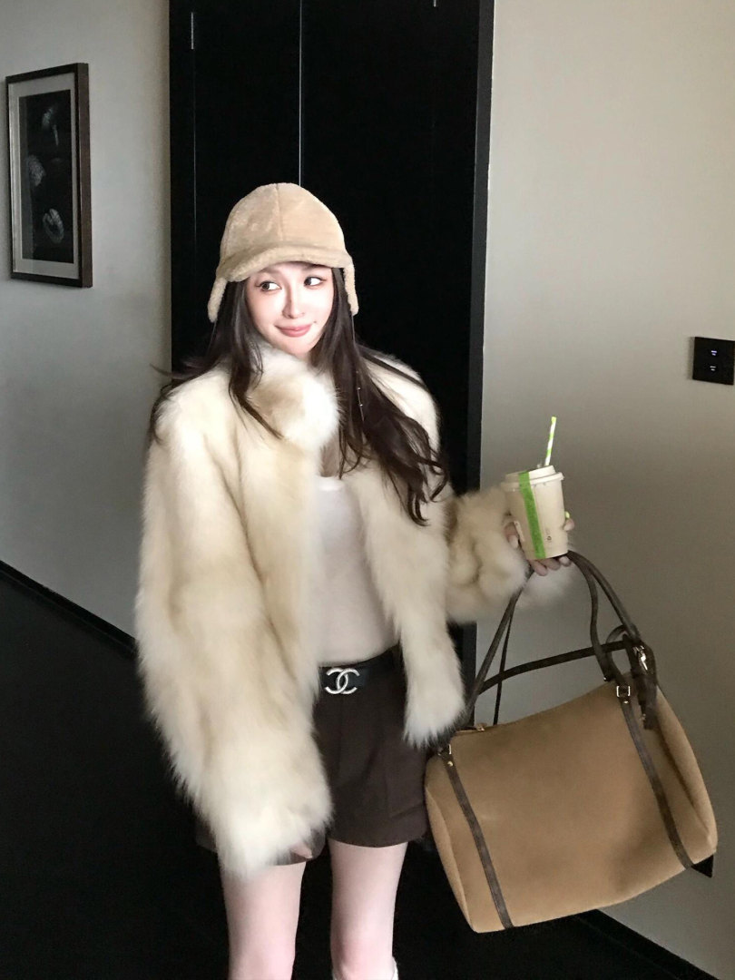 Woman modeling a cozy cream faux-fur coat with a knit hat, shorts, and large tan handbag