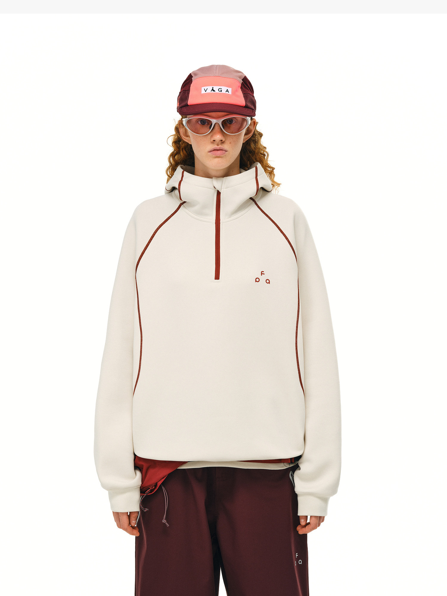 An individual with curly hair wears pink sunglasses, a red and white cap, maroon pants, and the EastLyst Retro-Core Half-Zip Hoodie—an off-white pullover with red trim—standing against a plain white background.